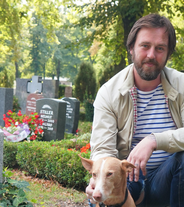 "37°: Mehr als Sarg und Service - Die neuen Bestatter*innen": Eric kniet vor Gräbern auf einem Friedhof, sein Hund sitzt neben ihm.