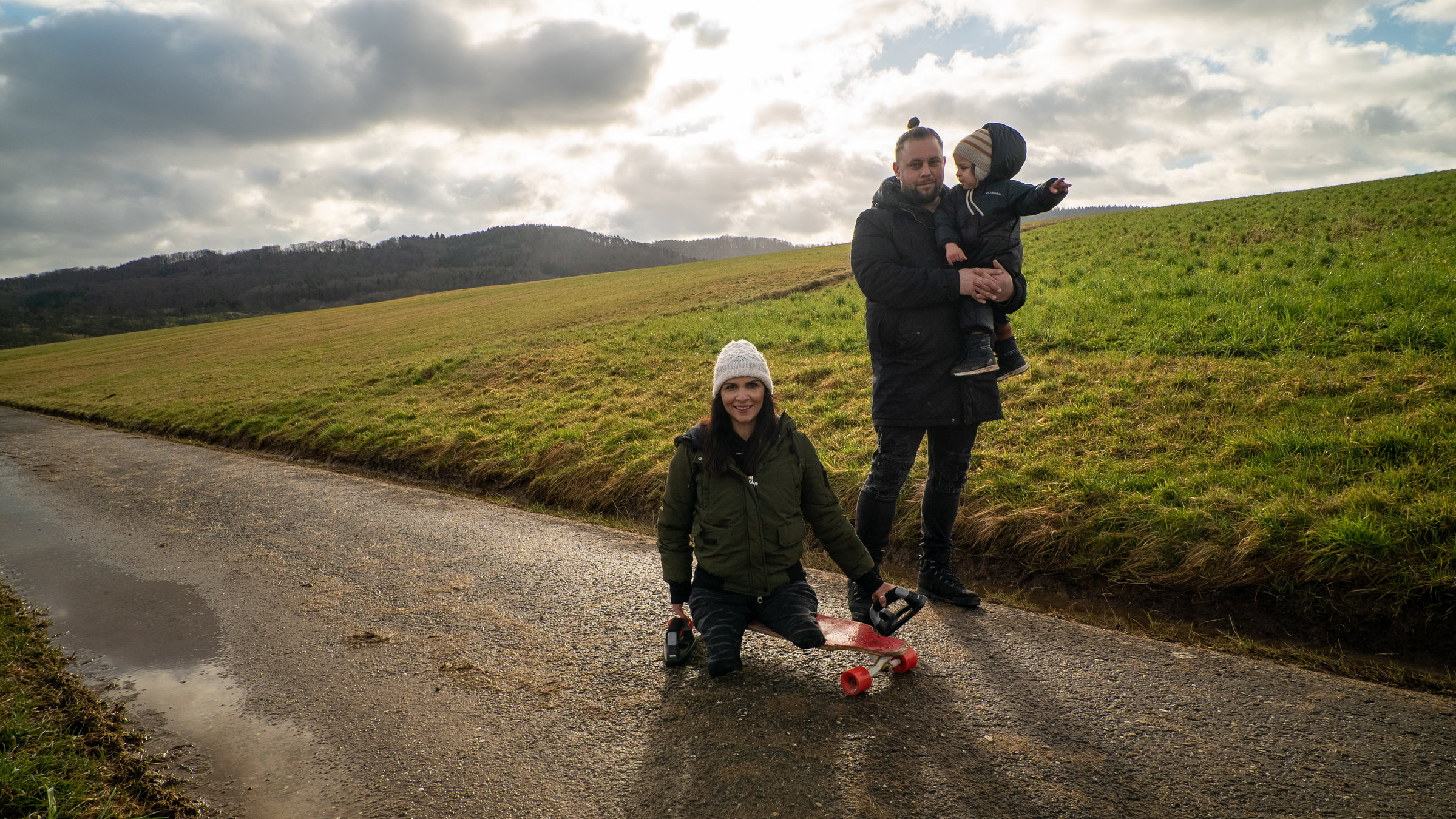 "Einfach Mensch! - Mitten im Leben": Hülya Marquardt (l.) sitzt auf einem Skateboard. Ihr Ehemann Dennis (m.) und ihr Sohn Rangi (r.) sind auch mit dabei. Sie sind auf einem Feldweg.