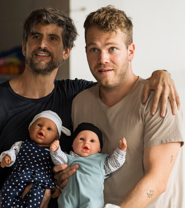 "Like a Loser: Ernsthaft?!": Guideon (Tom Beck) und Julian (Ben Münchow) stehen nebeneinander in einem Workshop-Raum. Beide halten lächelnd je eine Babypuppe in den Armen, die in niedliche Babystrampler gekleidet sind. Guideon wirkt ausgepowert, während Julian mit einem leicht überforderten, aber stolzen Gesichtsausdruck nach vorne schaut. Guideons Arm liegt locker auf Julians Schulter.