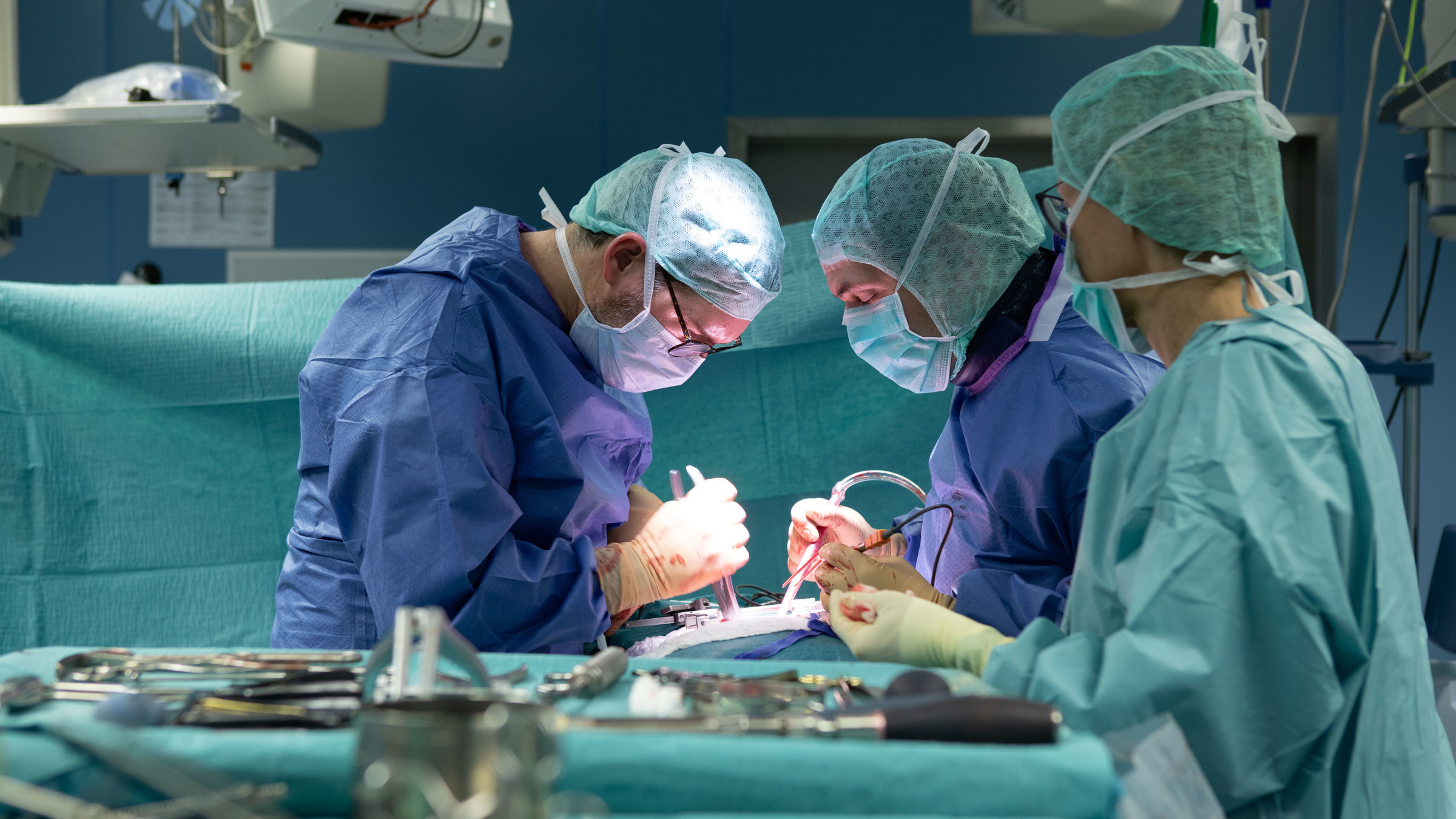 "plan b: Patientenwohl vor Profitstreben - Medizin im Sinne des Menschen":  Drei Menschen in OP-Kleidung operieren einen Patienten.