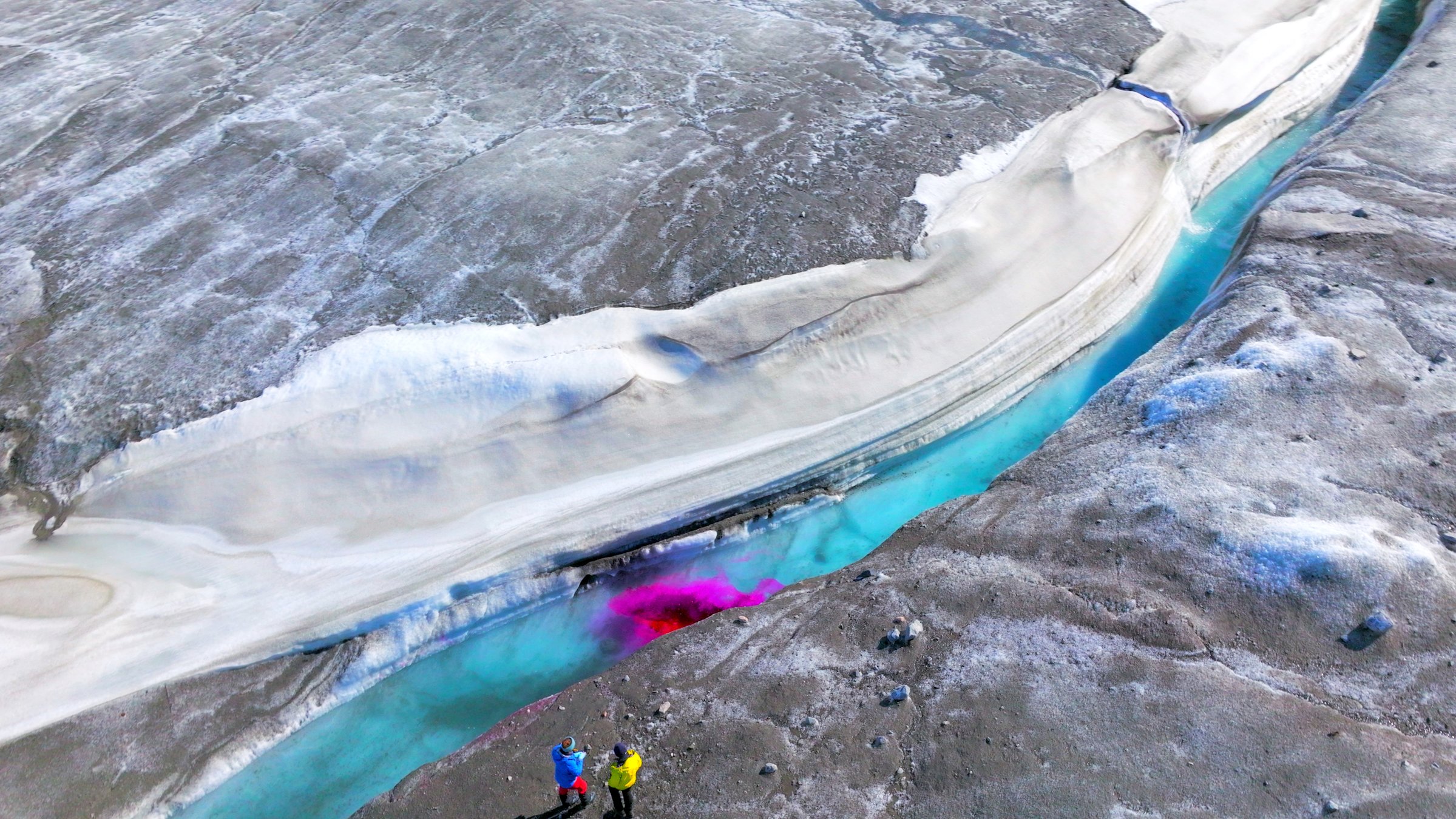 "Terra X:  Die Arktis – 66,5 Grad Nord - Eisige Schönheit": Blick von oben auf einen Fluss bei Kangerlussuaq in Grönland. Zwei Forscher stehen am Ufer. Sie haben zu Forschungszwecken ein pinkfarbenes Kontrastmittel in den Fluss geleitet.
