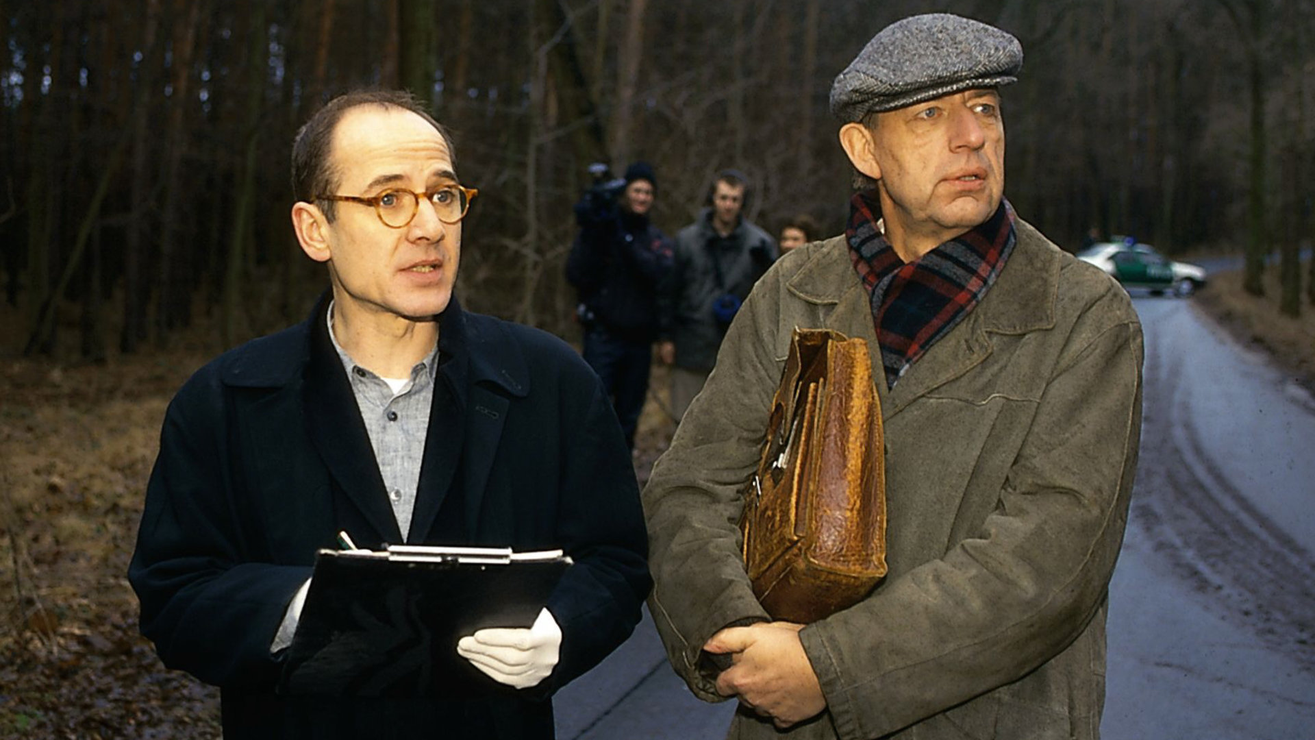 "Der letzte Zeuge - Der Weg zur Hölle": Kripochef Joe Hoffer (Jörg Gudzuhn) und Gerichtsmediziner Dr. Robert Kolmaar (Ulrich Mühe) stehen nebeneinander auf einer Straße im Wald und schauen aufmerksam in die gleiche Richtung.