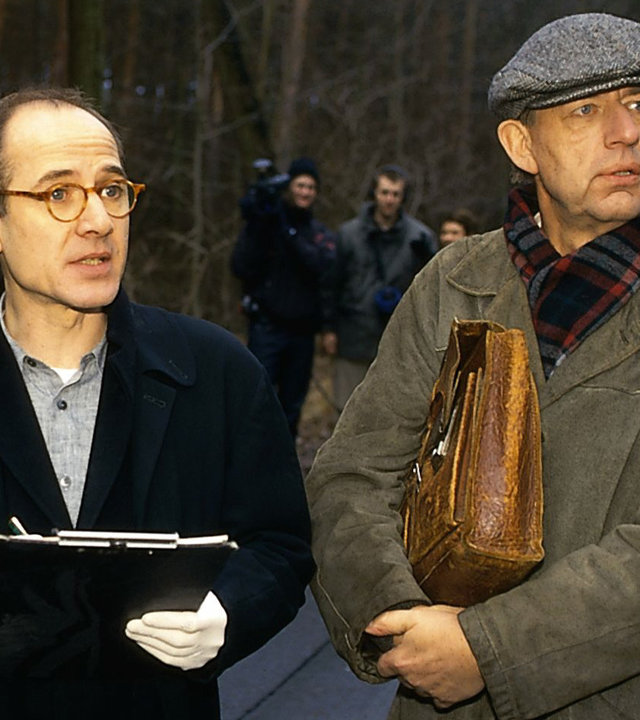 "Der letzte Zeuge - Der Weg zur Hölle": Kripochef Joe Hoffer (Jörg Gudzuhn) und Gerichtsmediziner Dr. Robert Kolmaar (Ulrich Mühe) stehen nebeneinander auf einer Straße im Wald und schauen aufmerksam in die gleiche Richtung.