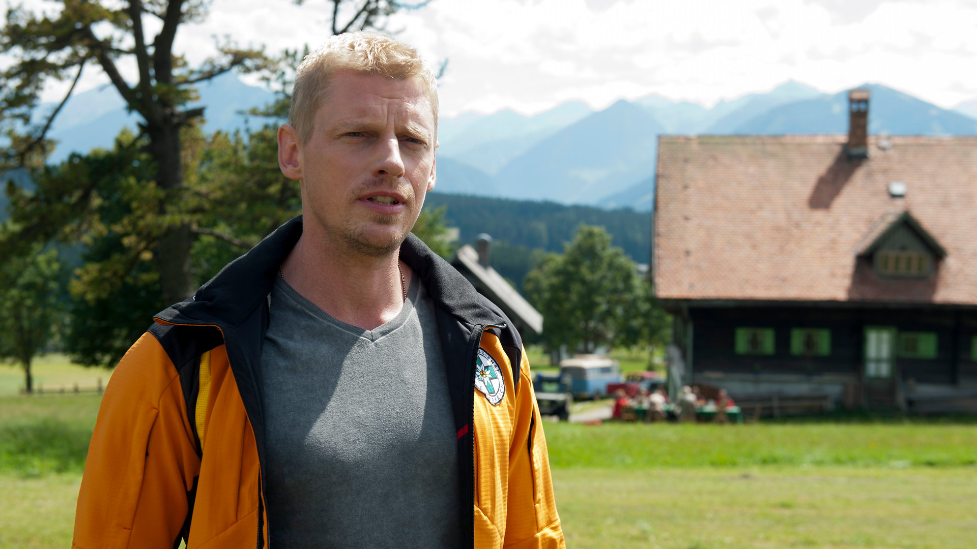 "Die Bergretter: Der Tote im Berg (1)": Andreas Marthaler (Martin Gruber) steht auf einer Wiese und blickt in die Ferne. Im Hintergrund steht ein Haus, vor dem an einem großen Tisch Menschen sitzen.