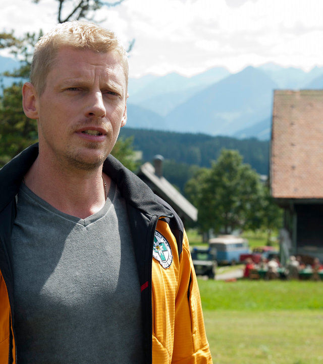 "Die Bergretter: Der Tote im Berg (1)": Andreas Marthaler (Martin Gruber) steht auf einer Wiese und blickt in die Ferne. Im Hintergrund steht ein Haus, vor dem an einem großen Tisch Menschen sitzen.