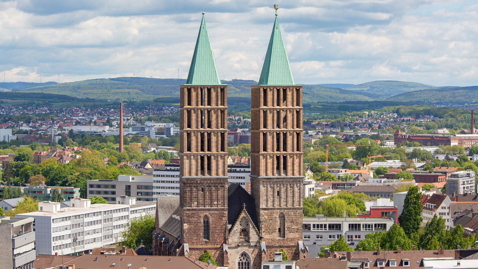 "Evangelischer Gottesdienst - Lauter Stimmen": Luftbild: Die Kirche St. Martin steht inmitten von Kassel. Ihre zwei eckigen Türme mit grünen Spitzen ragen hoch über die Stadt. Im Hintergrund liegen die sanften Hügel des Habichtswälder Berglandes.