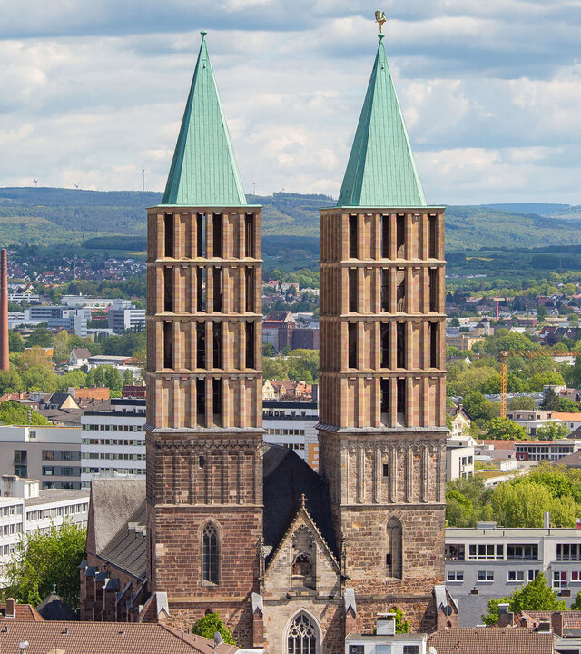 "Evangelischer Gottesdienst - Lauter Stimmen": Luftbild: Die Kirche St. Martin steht inmitten von Kassel. Ihre zwei eckigen Türme mit grünen Spitzen ragen hoch über die Stadt. Im Hintergrund liegen die sanften Hügel des Habichtswälder Berglandes.