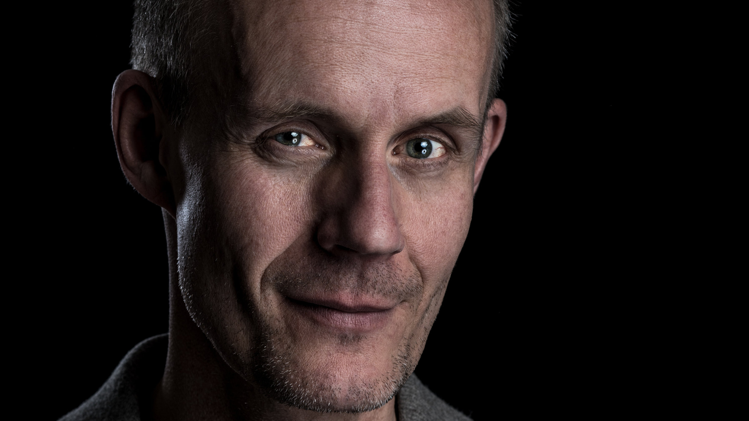 "Max Uthoff: Moskauer Hunde": Portrait von Max Uthoff. Er trägt ein graues Shirt und einen grauen Blazer und steht vor einem schwarzen Hintergrund.