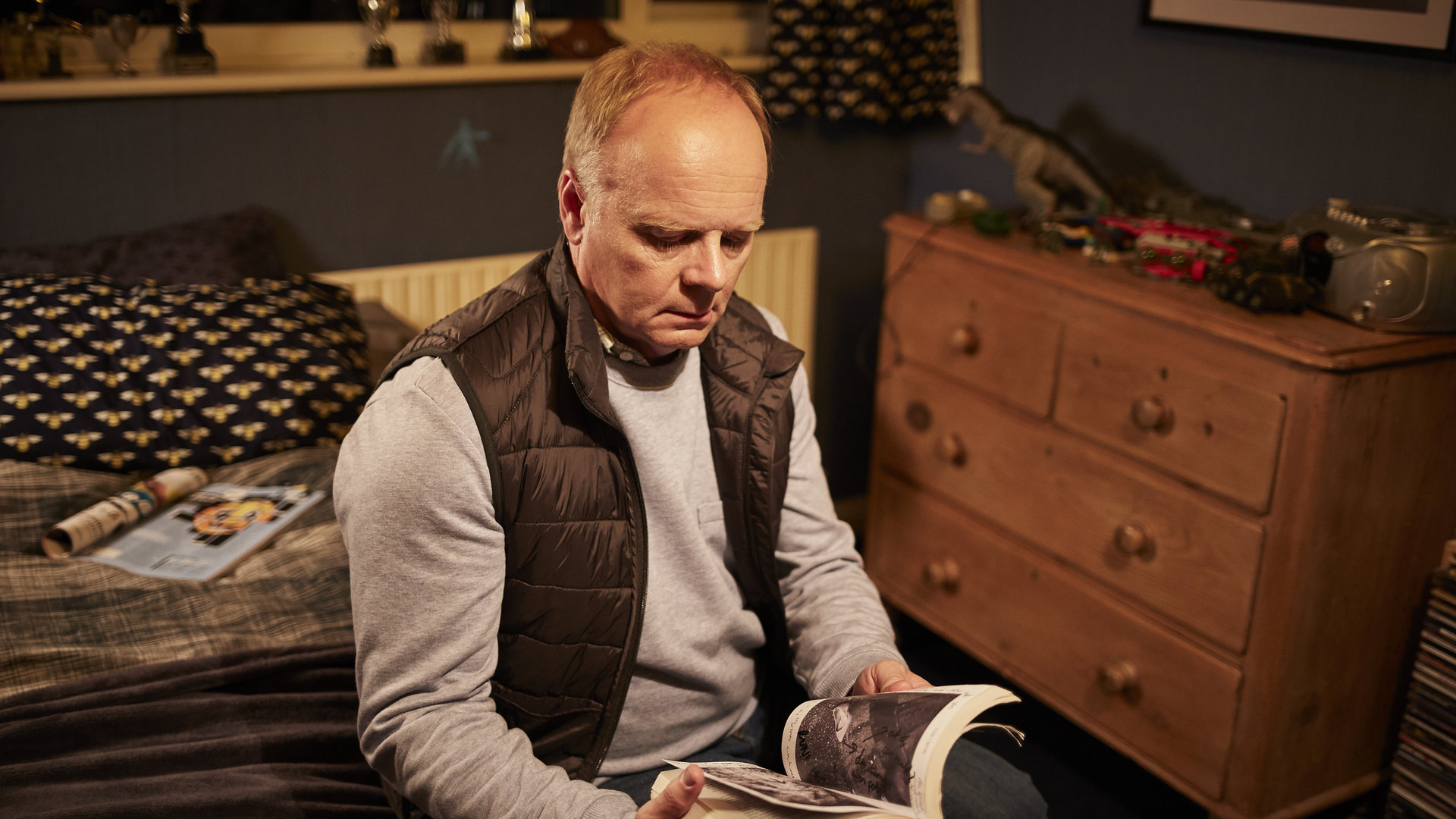 "Safe House - Verdacht": Simone Duke (Jason Watkins) sitzt auf einem Bett in einem Schlafzimmer und schaut in ein aufgeschlagenes Buch, das er in den Händen hält.