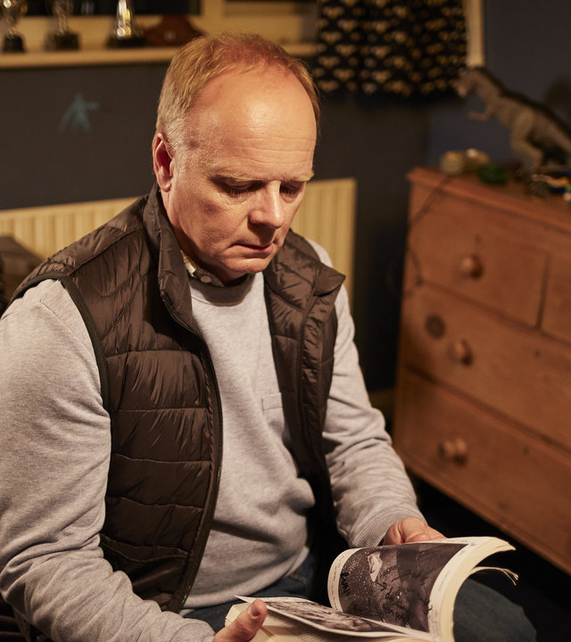 "Safe House - Verdacht": Simone Duke (Jason Watkins) sitzt auf einem Bett in einem Schlafzimmer und schaut in ein aufgeschlagenes Buch, das er in den Händen hält.