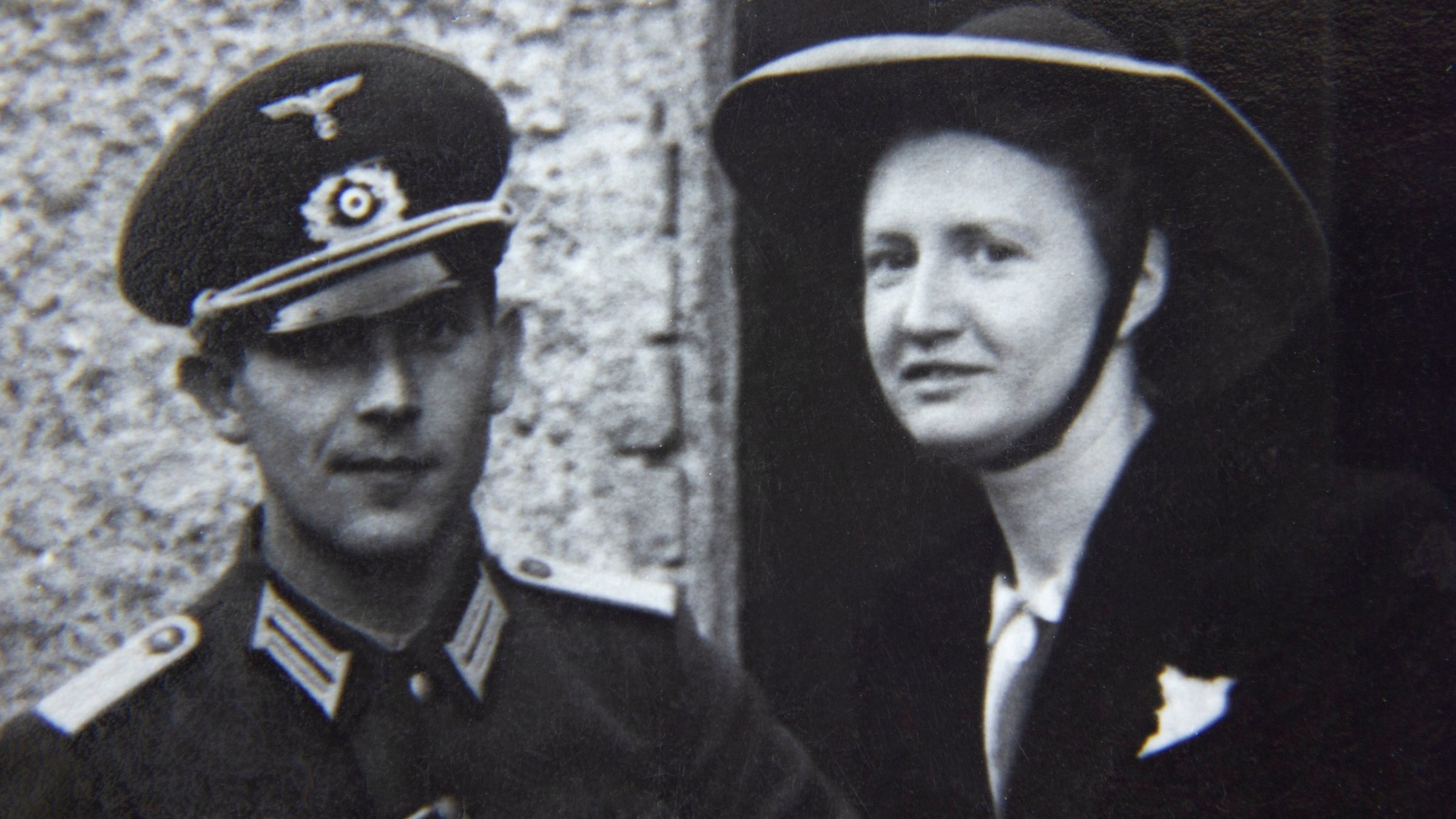"Der Sog des Krieges": Historisches Schwarzweiß-Foto eines jungen Paares, das vor einem Haus steht. Der Mann trägt militärische Uniform mit Abzeichen des Dritten Reichs und Schirmmütze, die Frau einen breitrandigen Hut mit Kinnband und eine dunkle, elegante Jacke. Beide schauen lächelnd in die Kamera.