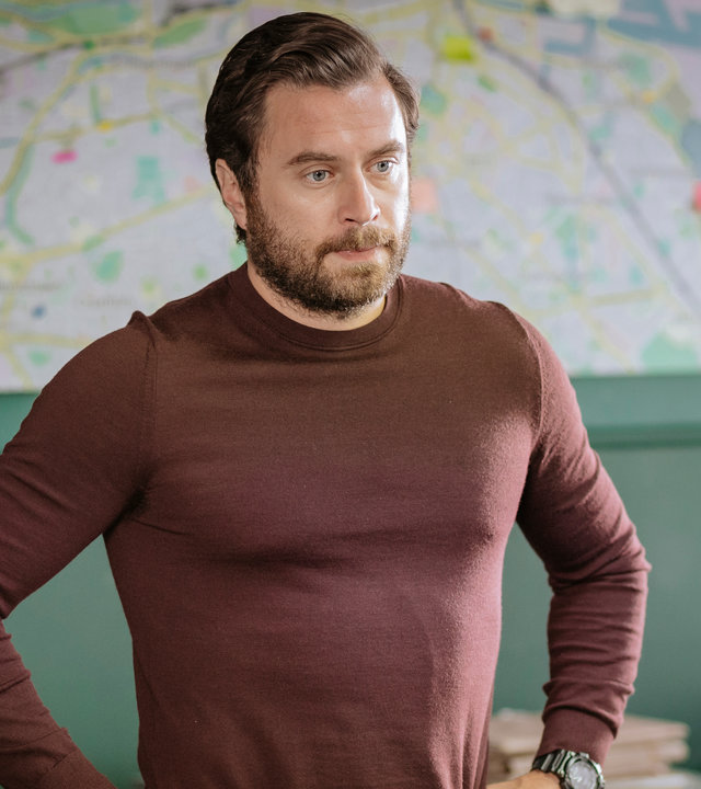 "Harry Wild - Mörderjagd in Dublin: Wenn Happy nicht happy ist, ist niemand happy": Charlie Wild (Kevin J. Ryan) steht in einem Büro, seine Arme in die Hüften gestützt. Er schaut ernst.