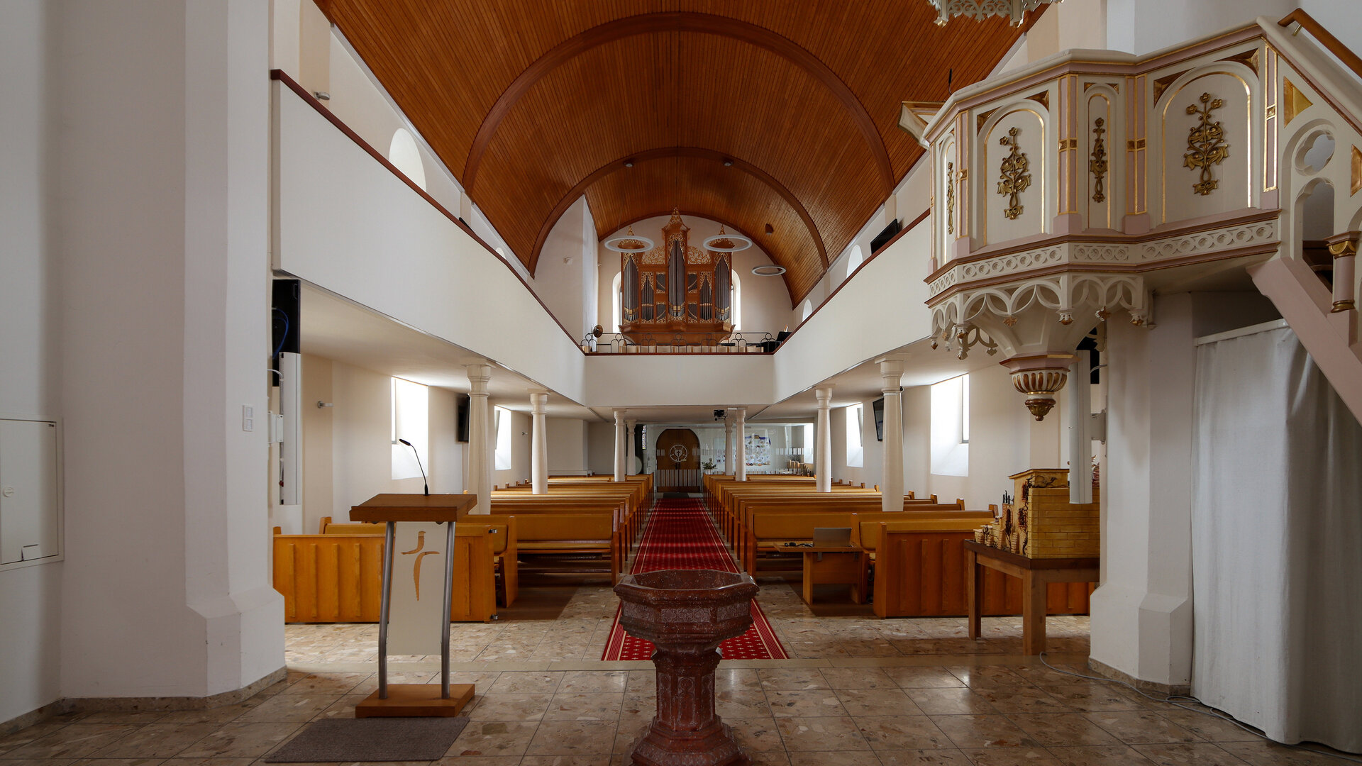 "Evangelischer Gottesdienst - Ja, ich bin getauft!": Innenansicht der Evangelischen Kirche im oberösterreichischen Rutzenmoos. In der vorderen Mitte steht das alte Taufbecken, an der Seite befindet sich die elfenbeinweiße, goldverzierte Kanzel. Der Blick reicht durch das Kirchenschiff bis hinter zum Altar und der sich darüber befindenden Orgel. Kirchgestühl und Decke sind schlicht in hellem Holz gehalten.