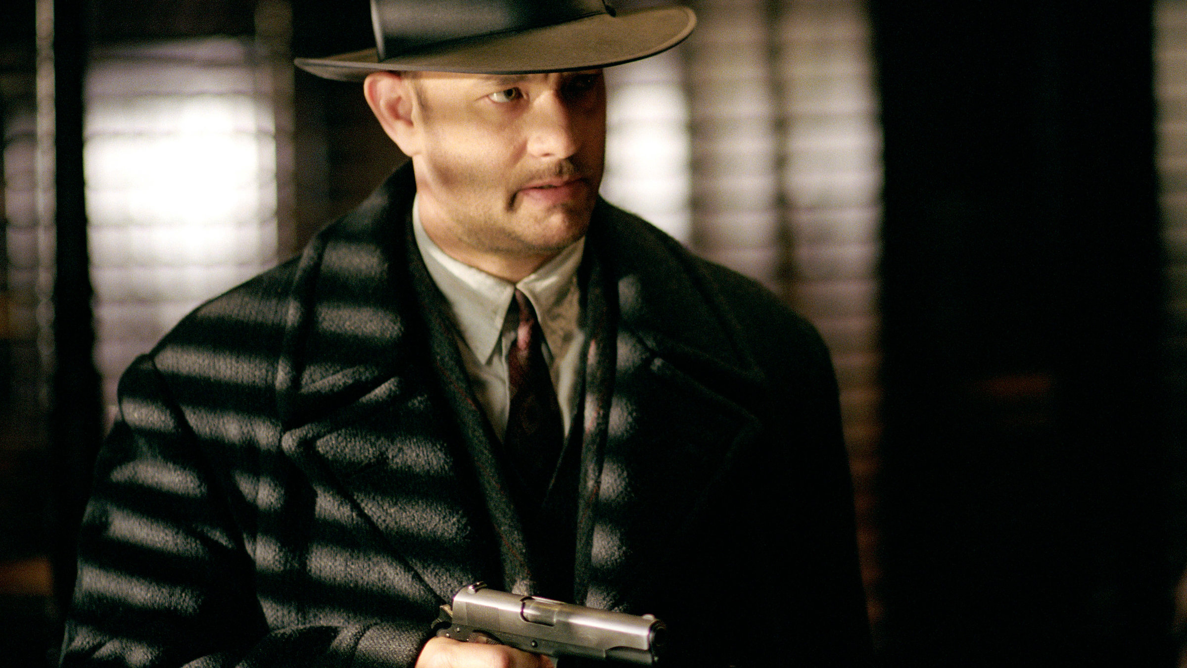 "Road to Perdition": Elegant gekleidet mit Mantel, Krawatte und Hut, in der rechten Hand eine Pistole, bewegt sich Michael Sullivan (Tom Hanks) vorsichtig in einem dunklen Raum, in den wenig Licht fällt.
