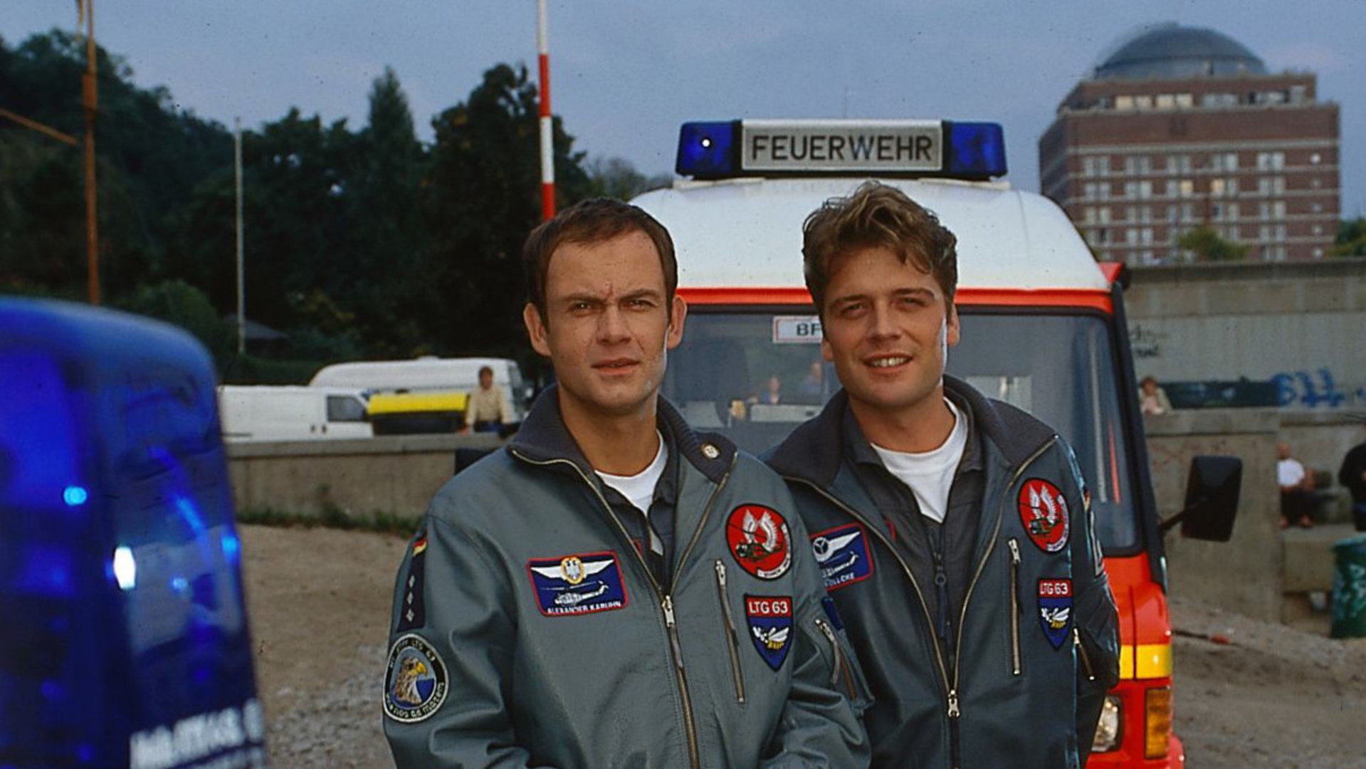 "Die Rettungsflieger - Auf Gedeih und Verderb": Pilot Alex (Matthias Leja) und Wollcke (Oliver Hörner) stehen nebeneinander auf einer Snadfläche vor einem Rettungswagen der Feuerwehr.