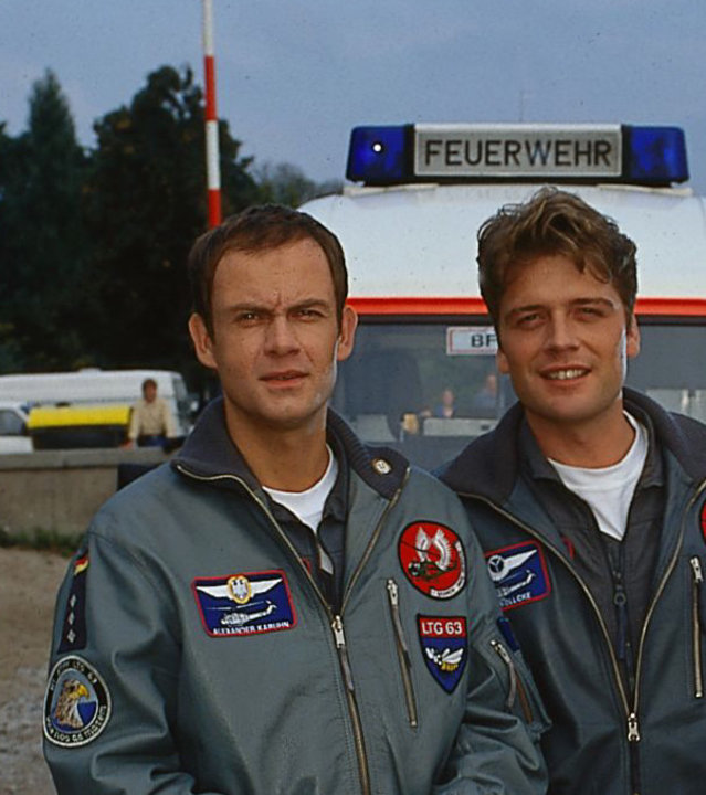 "Die Rettungsflieger - Auf Gedeih und Verderb": Pilot Alex (Matthias Leja) und Wollcke (Oliver Hörner) stehen nebeneinander auf einer Snadfläche vor einem Rettungswagen der Feuerwehr.