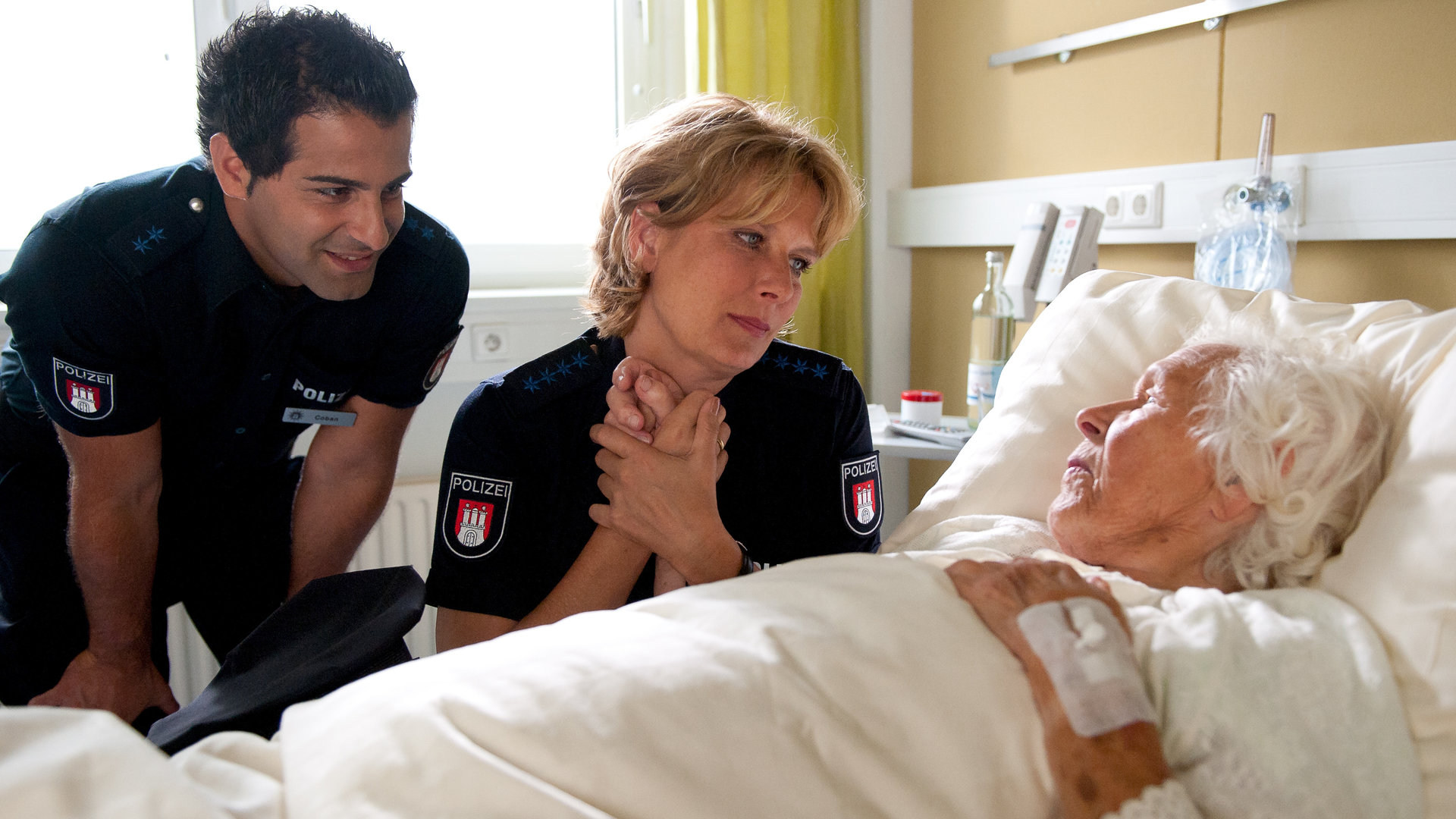 "Notruf Hafenkante - Schutzengel": Elsa (Renate Delfs) liegt in einem Krankenhausbett. Claudia (Janette Rauch), die neben ihr sitzt, hält liebevoll ihre Hand und sieht Elsa an. Tarik (Serhat Cokgezen), der ebenfalls neben dem Bett steht, lächelt die alte Dame aufmunternd an.