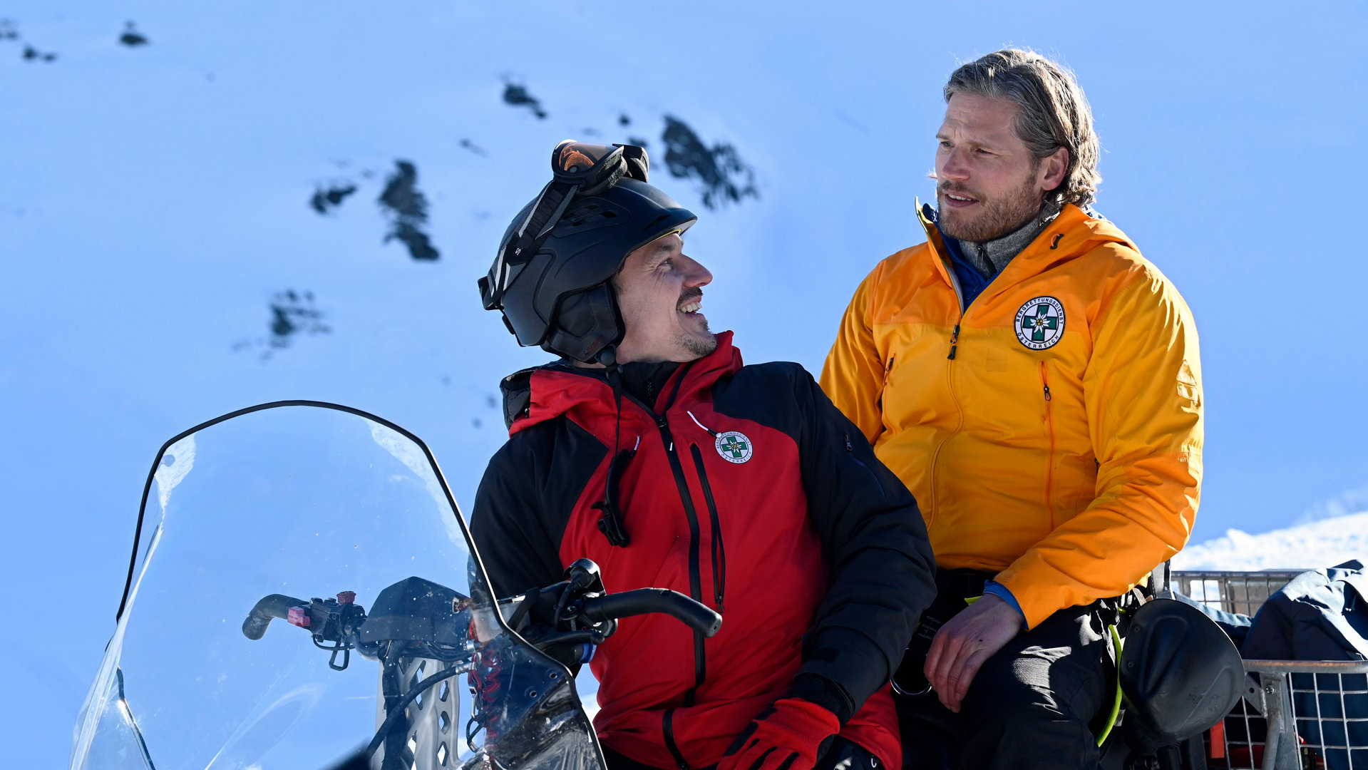 "Die Bergretter: Giftiges Erbe": Vor einer Schneekulisse sitzt Tobi (Markus Brandl) auf einem Ski-doo und blickt lachend über die Schulter nach hinten zu Markus (Sebastian Ströbel).