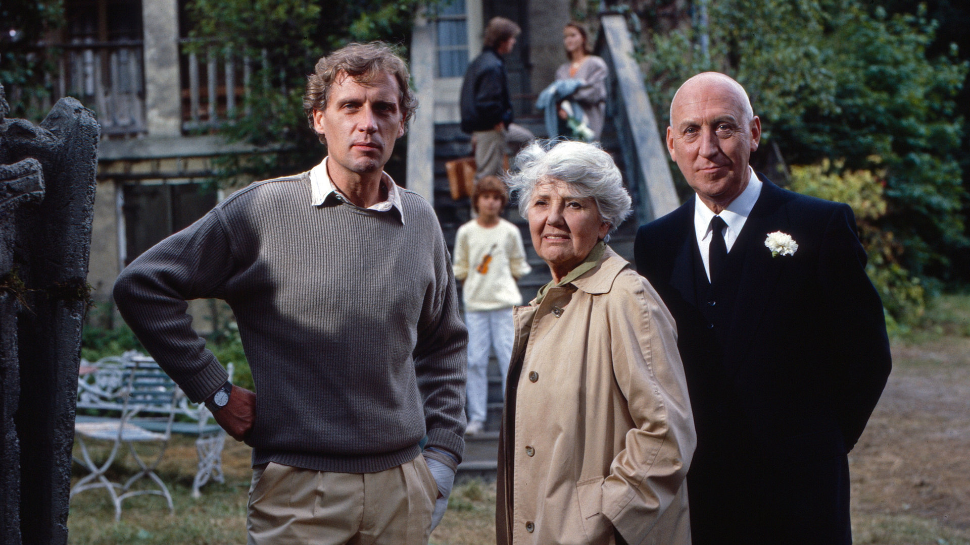 "Oliver Maass - Das Spiel mit der Zaubergeige": Michael Maass (Robert Atzorn), Louise Maass (Jane Tilden) und Eickelberg (Karl Lieffen) stehen nebeneinander in einem Vorgarten und schauen ernst in die Ferne.