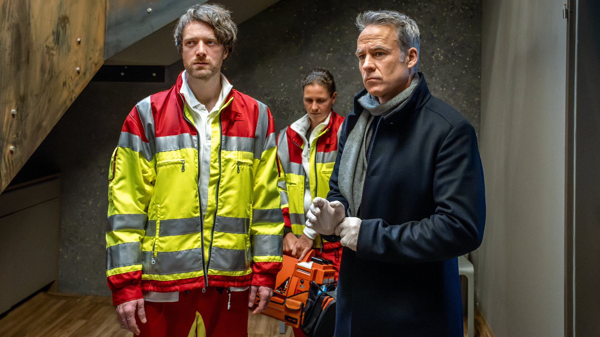 "SOKO Leipzig - Bittere Medizin": Benni Maybach (Maximilian Klas) und Jan Maybach (Marco Girnth) stehen am Tatort und schauen ernst. Im Hintergrund steht eine Rettungssanitäterin (Komparsin).