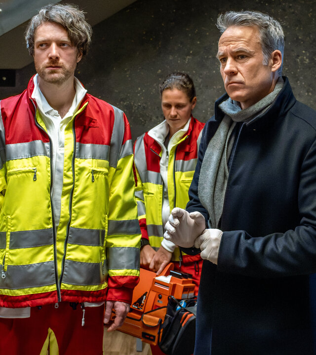 "SOKO Leipzig - Bittere Medizin": Benni Maybach (Maximilian Klas) und Jan Maybach (Marco Girnth) stehen am Tatort und schauen ernst. Im Hintergrund steht eine Rettungssanitäterin (Komparsin).