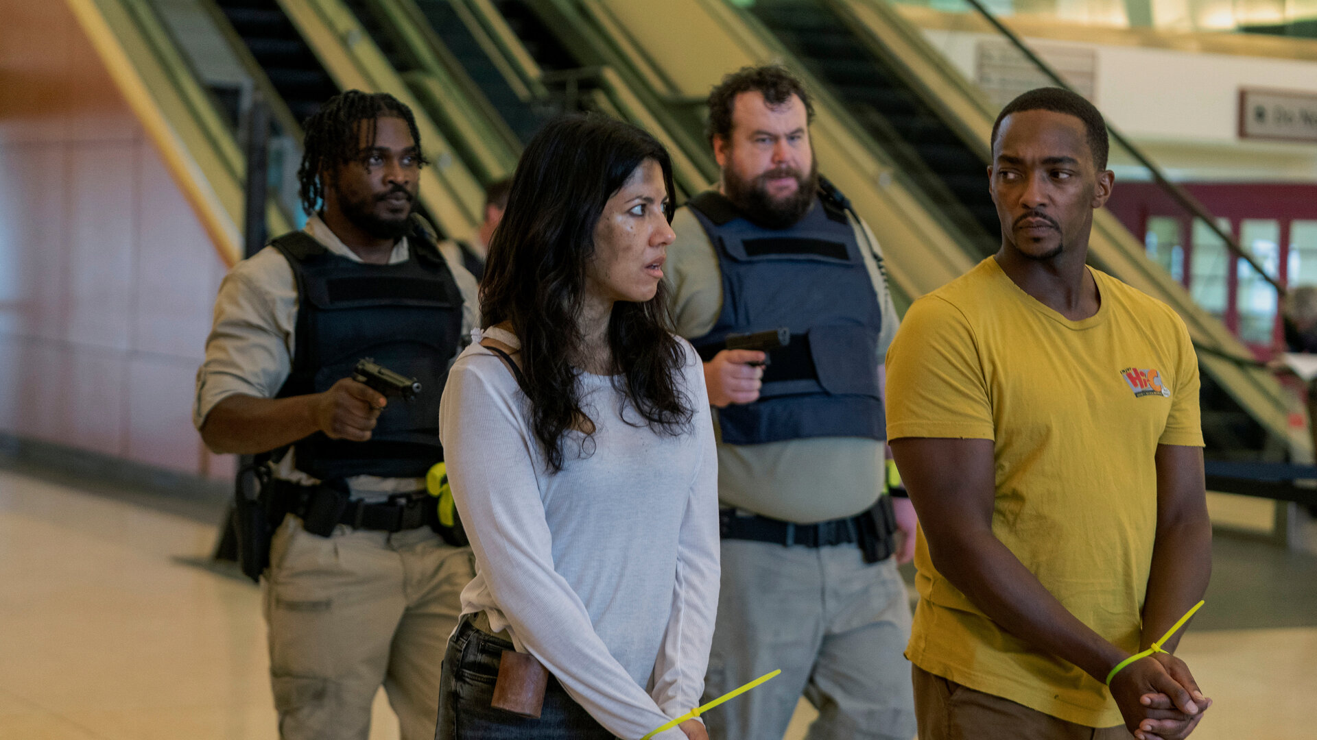 "Twisted Metal - NTHLAW1": John (Anthony Mackie) und Quiet (Stephanie Beatriz) stehen mit gefesselten Händen in einem öffentlichen Gebäude. Dicht hinter ihnen halten Stu (Mike Mitchell) und Mike (Tahj Vaughans) mit schusssicheren Westen jeweils eine Waffe auf sie gerichtet.