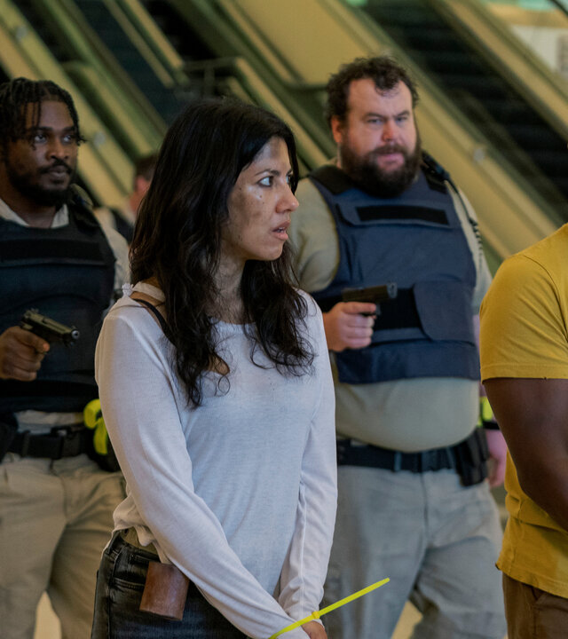 "Twisted Metal - NTHLAW1": John (Anthony Mackie) und Quiet (Stephanie Beatriz) stehen mit gefesselten Händen in einem öffentlichen Gebäude. Dicht hinter ihnen halten Stu (Mike Mitchell) und Mike (Tahj Vaughans) mit schusssicheren Westen jeweils eine Waffe auf sie gerichtet.