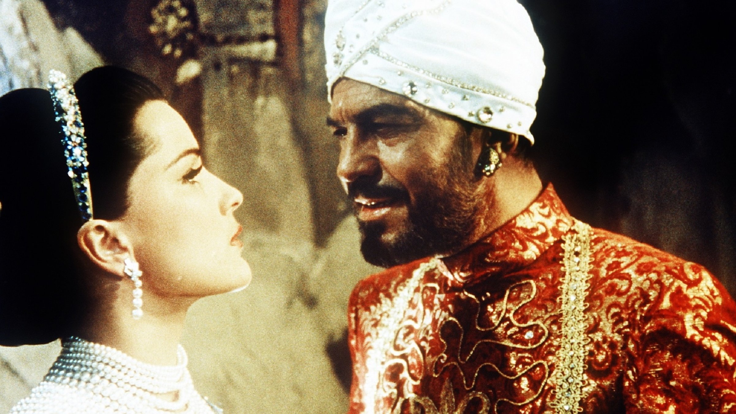 "Das indische Grabmal": Ein dunkelhäutiger, bärtiger Mann mit weißem Turban nähert sich mit spöttischem Blick von rechts einer festlich hell gekleideten Frau mit halblangem braunem Haar, die mit zahlreichen prunkvollen Perlenketten geschmückt ist und ihn eingeschüchtert anblickt.