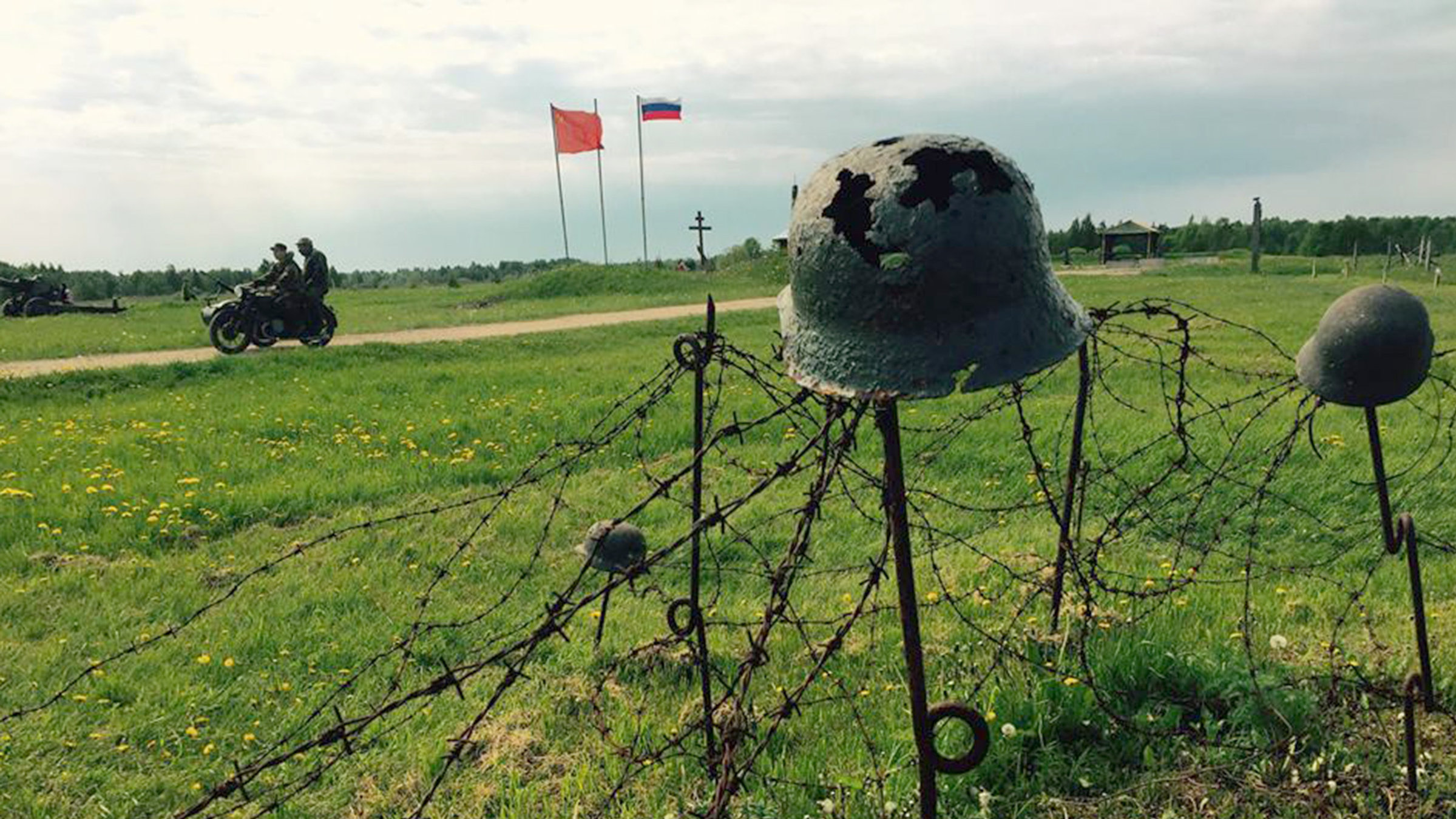 "Grenzland - Vom Baltikum zur Akropolis - Von Estland bis in den Westen der Ukraine": In Vordergrund sitzt ein rostiger Stahlhelm auf einem Stab, im Hintergrund fährt ein Motorrad einen schmalen Weg entlang, dahinter wehen Fahnen.