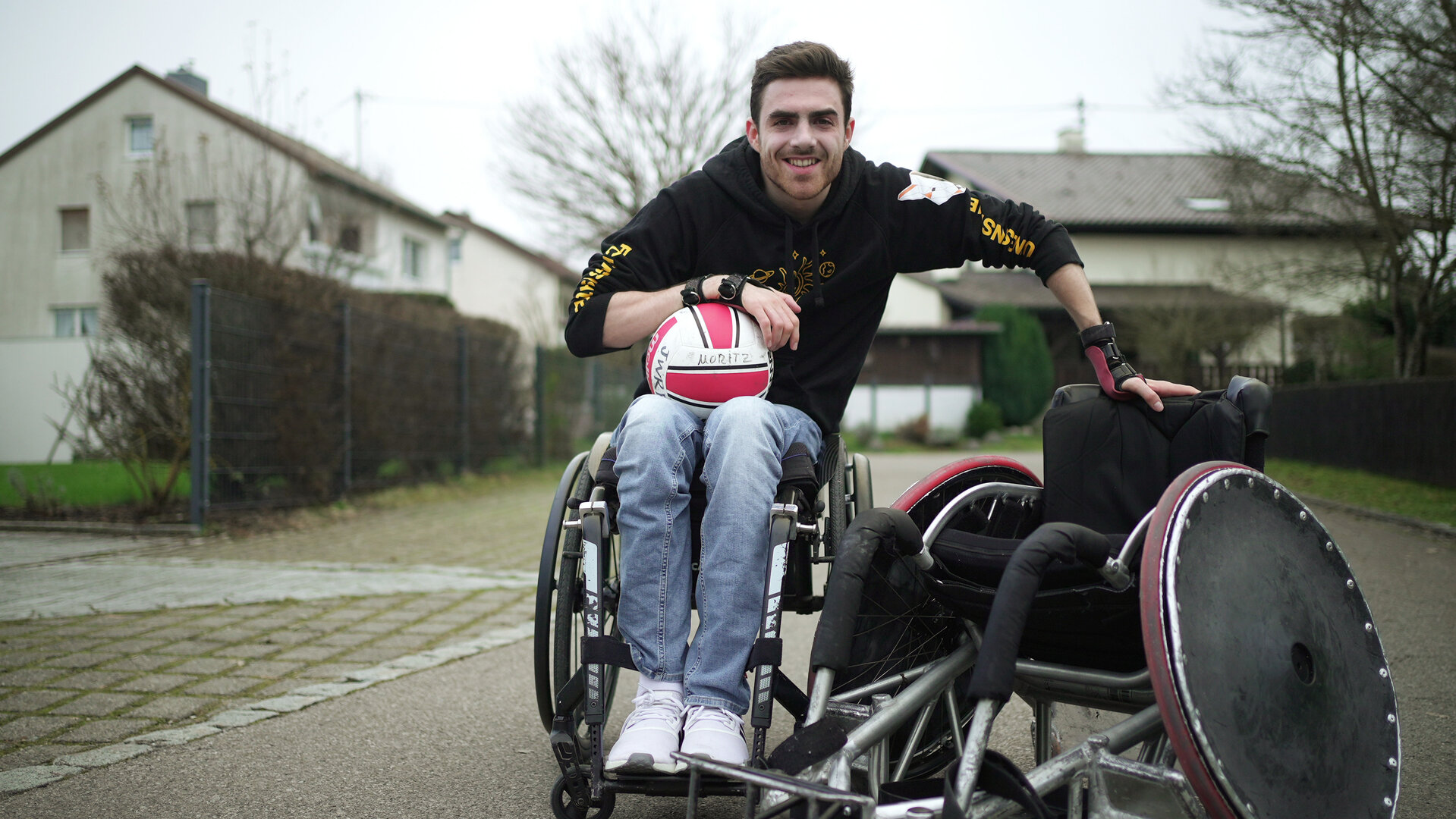 "einfach Mensch - Moritz Brückner: Für mehr Inklusion in Deutschland": Moritz Brückner lehnt sich an einen Rugby-Rollstuhl und hat einen Ball auf dem Schoß.