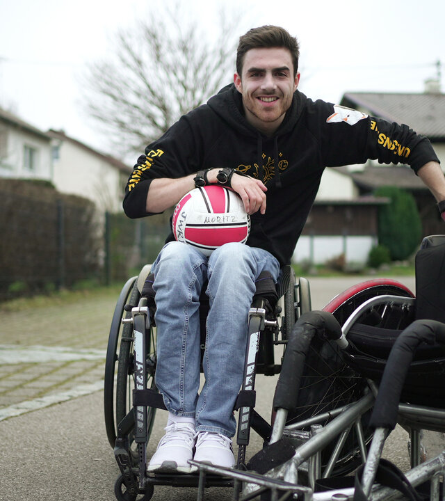 "einfach Mensch - Moritz Brückner: Für mehr Inklusion in Deutschland": Moritz Brückner lehnt sich an einen Rugby-Rollstuhl und hat einen Ball auf dem Schoß.