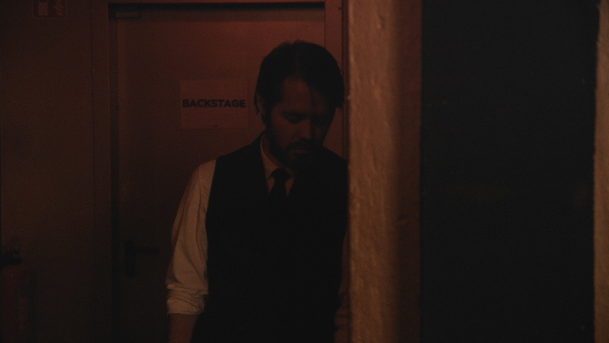"Wie ein Fremder - Eine deutsche Popmusik-Geschichte - Shine": Junger, bärtiger Mann halbnah, der mit gesenktem Kopf an einer Wand lehnt. Im Hintergrund eine Tür mit der Aufschrift „Backstage“.