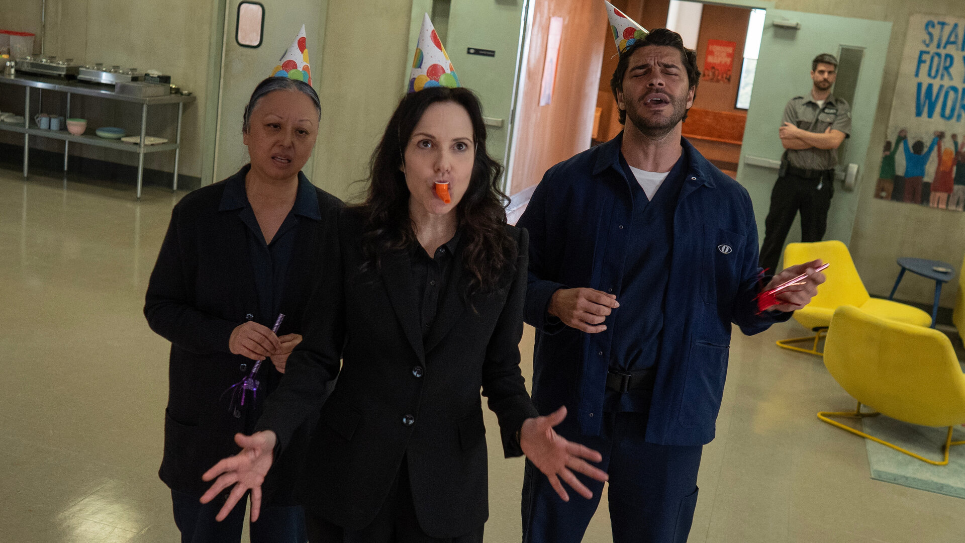 "Das Institut - Jetzt oder nie": Maureen (Jane Luk), Ms. Sigsby (Mary-Louise Parker) und Tony (Jason Diaz) stehen in einem großen Raum und singen. Alle tragen jeweils ein buntes Partyhütchen auf dem Kopf. Sigsby hat zudem ein Plastikkazoo im Mund. An der geöffneten Tür steht ein Security-Mitarbeiter (Xander Martel).