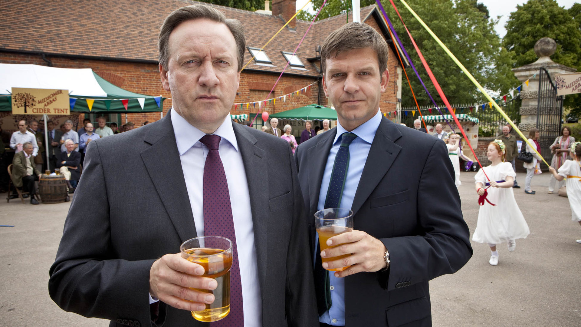 "Inspector Barnaby - Das Biest muss sterben": DCI John Barnaby (Neil Dudgeon), DS Ben Jones (Jason Hughes) auf einem Dorffest. Beide, wie immer in Anzug und Krawatte, halten jeweils ein Getränk in der Hand.