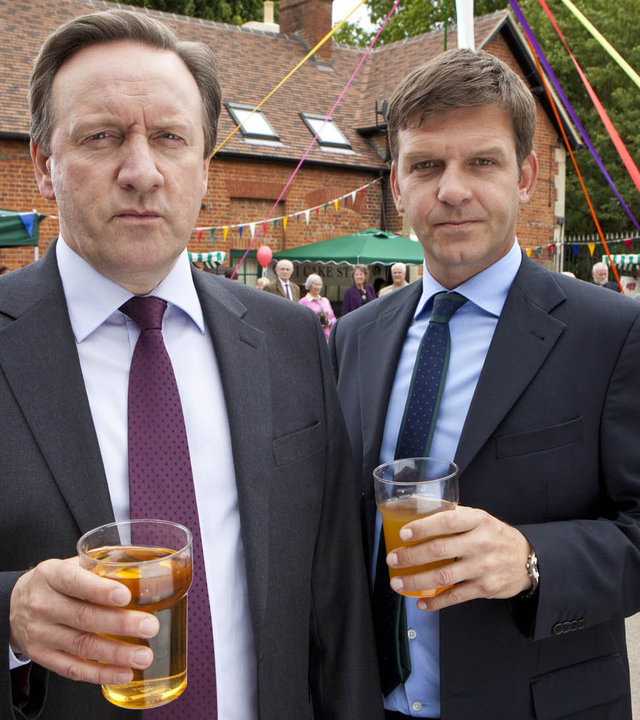 "Inspector Barnaby - Das Biest muss sterben": DCI John Barnaby (Neil Dudgeon), DS Ben Jones (Jason Hughes) auf einem Dorffest. Beide, wie immer in Anzug und Krawatte, halten jeweils ein Getränk in der Hand.