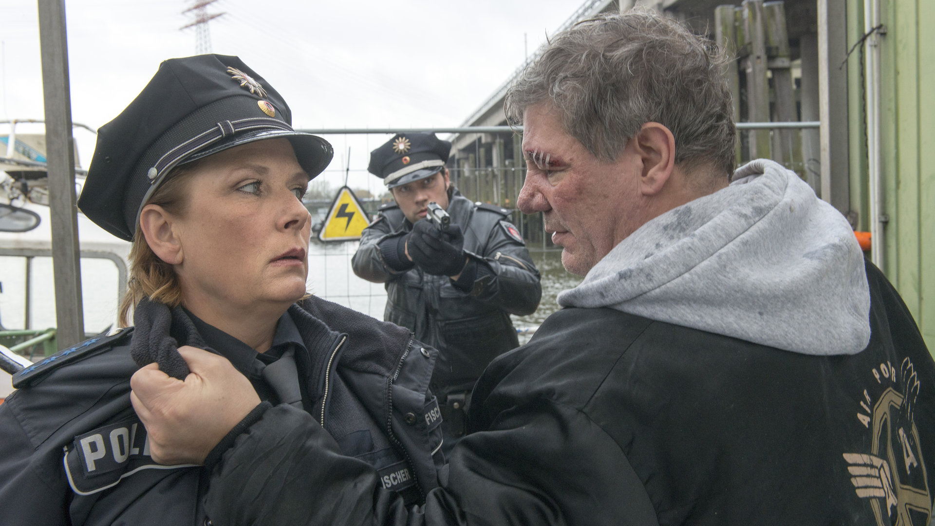"Notruf Hafenkante - Lucky Punch": Mitschke (Claude-Oliver Rudolph) packt Claudia (Janette Rauch) am Kragen und wirkt sehr aggressiv. Im Hintergrund steht Mattes (Matthias Schloo) mit gezogener Waffe.
