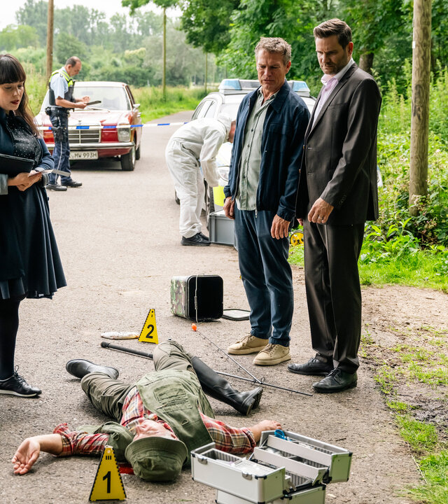 "SOKO Köln - Tödliche Harmonie": Matti Wagner (Pierre Besson) und Jonas Fischer (Lukas Piloty) stehen an einem Tatort auf einer Straße im Grünen gegenüber von Dr. Pilar Westphal (Yulia Yáñez Schmidt). Sie blicken nach unten auf einen Mann (Komparse), der vor ihnen auf dem Rücken mit ausgestreckten und verdrehten Gliedmaßen liegt. Die Rechtsmedizinerin hält ein Klemmbrett im Arm und sieht ebenfalls zu dem Toten. Neben ihr steht ein schwarzer Rollkoffer. Rechts auf einem Gehweg kniet eine Mitarbeiterin (Komparsin) der Spurensicherung und notiert etwas in einer Mappe. Im Hintergrund stehen zwei Polizeiautos und ein roter Pkw. Daneben steht ein weiterer Mitarbeiter (Komparse) der Spurensicherung und ein uniformierter Polizist (Komparse).