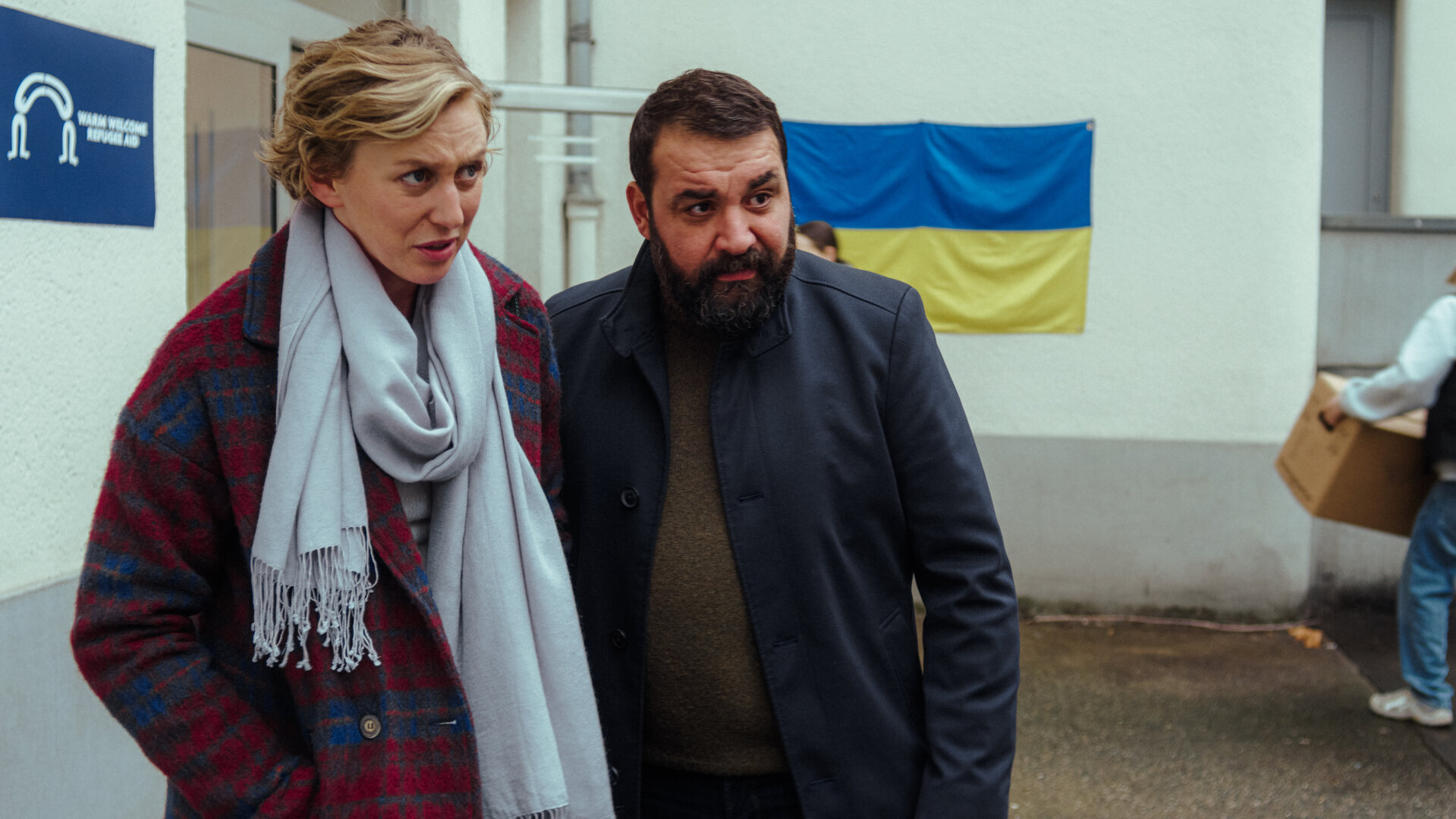 "Uncivilized - Ukraine": Karla (Franziska Machens) und Eren (Sahin Eryilmaz) stehen vor der Flüchtlingsunterkunft, während im Hintergrund eine Person einen Karton trägt.