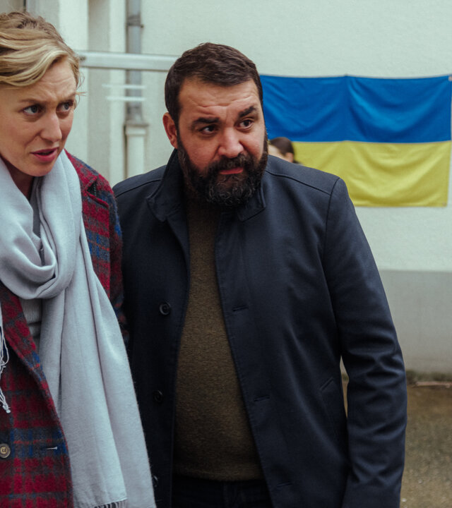 "Uncivilized - Ukraine": Karla (Franziska Machens) und Eren (Sahin Eryilmaz) stehen vor der Flüchtlingsunterkunft, während im Hintergrund eine Person einen Karton trägt.