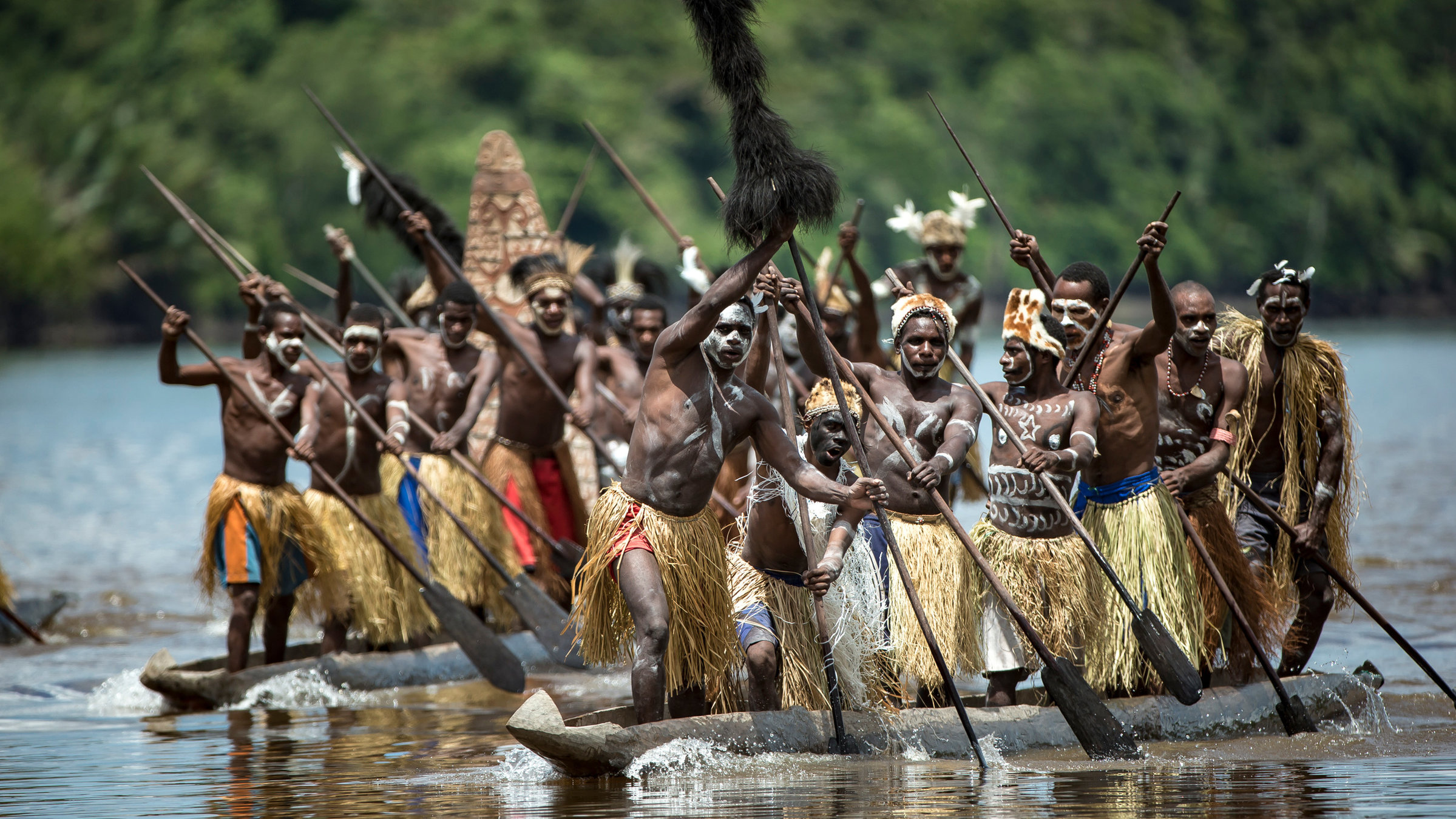 "Schutzprojekt Erde": Viele schwarze Männer mit Kriegsbemalung, stehend auf mehreren einfachen Holzbooten, die mit Paddeln bedient werden.