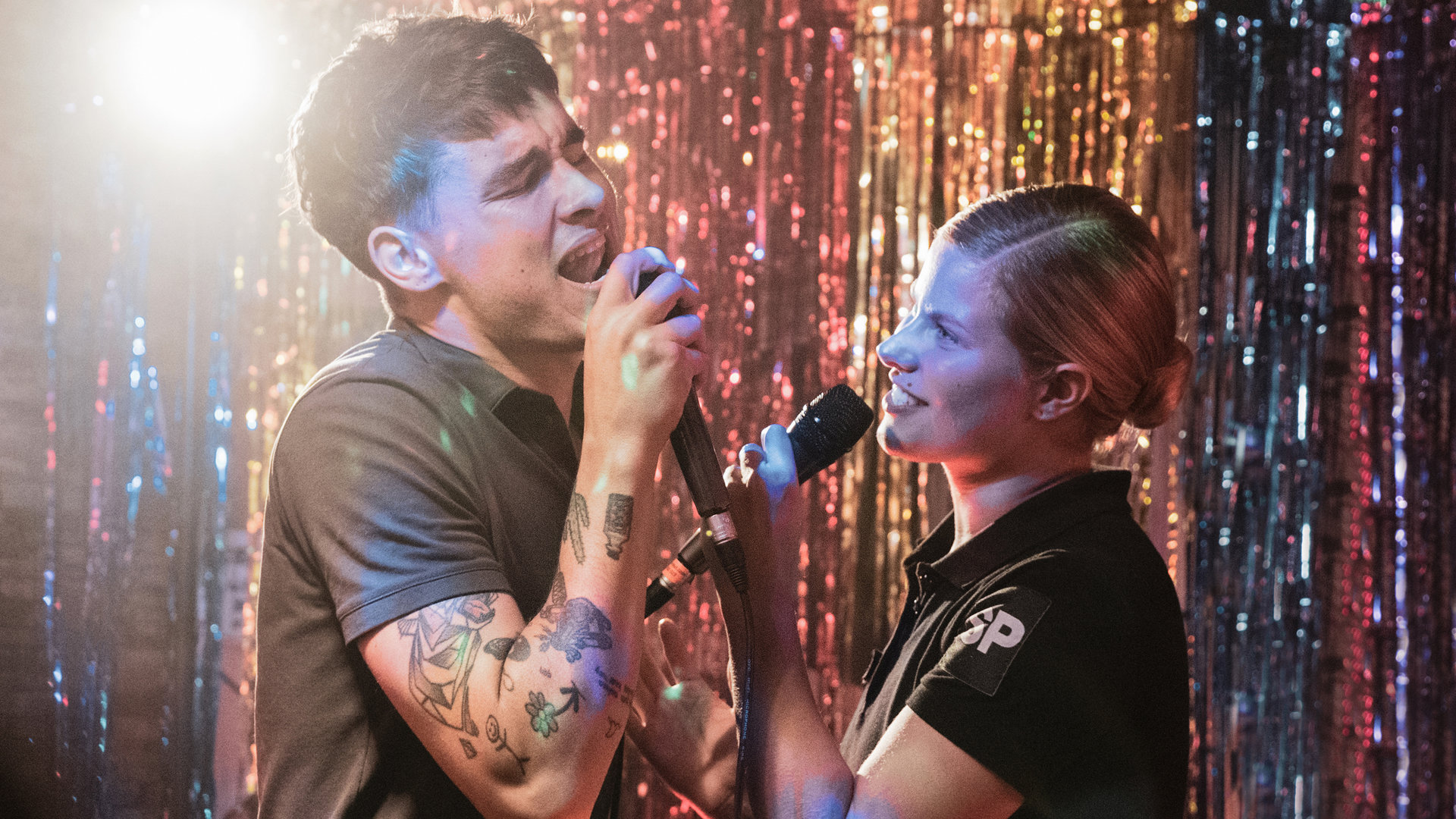 "Californian Commando - Auf der Flucht": Van (Kian Lawley) und Minttu (Wenla Reimaluoto) stehen gemeinsam auf einer Bühne und singen leidenschaftlich in die Mikrophone in ihren Händen.