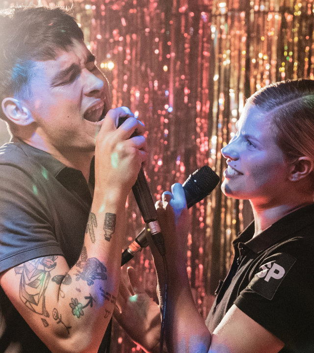 "Californian Commando - Auf der Flucht": Van (Kian Lawley) und Minttu (Wenla Reimaluoto) stehen gemeinsam auf einer Bühne und singen leidenschaftlich in die Mikrophone in ihren Händen.