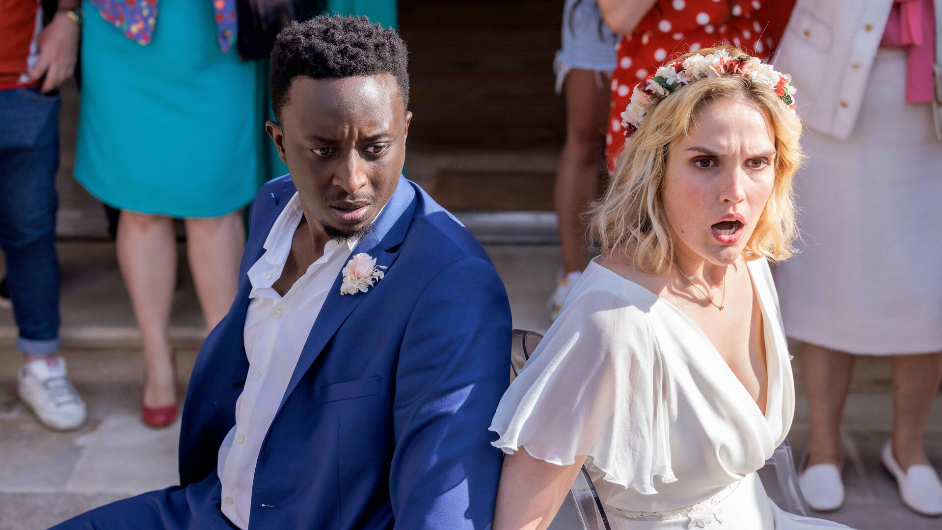 "Tiny Wedding - Unsere mega kleine Hochzeit": Halbnahe, leichte Aufsicht: Max (Ahmed Sylla) und Lou (Camille Lou) sitzen in ihren Hochzeitsoutfits mit schockierten Gesichtern Rücken an Rücken an Stühle gefesselt vor einer Feiergesellschaft. Diese ist nur hüftabwärts im Bild zu sehen.