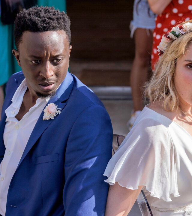 "Tiny Wedding - Unsere mega kleine Hochzeit": Halbnahe, leichte Aufsicht: Max (Ahmed Sylla) und Lou (Camille Lou) sitzen in ihren Hochzeitsoutfits mit schockierten Gesichtern Rücken an Rücken an Stühle gefesselt vor einer Feiergesellschaft. Diese ist nur hüftabwärts im Bild zu sehen.