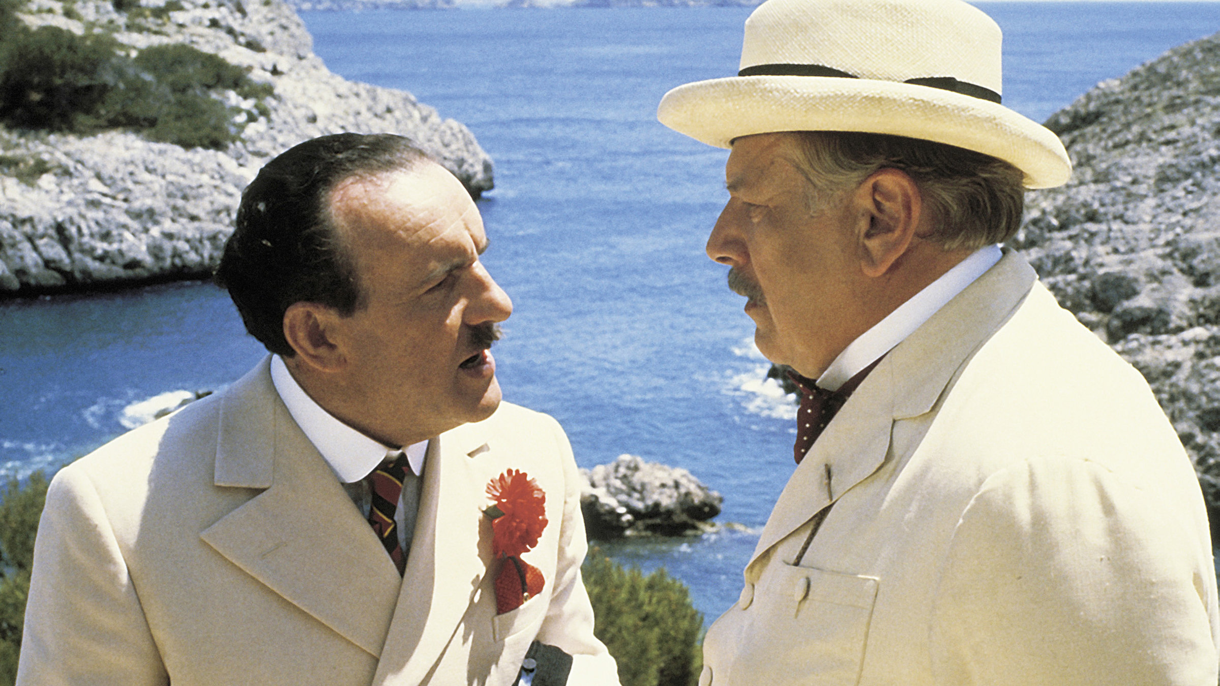 „Das Böse unter der Sonne“: Hercule Poirot (Peter Ustinov) mit Hut und Sir Horace Blatt (Colin Blakely, links) befinden sich, elegant-sommerlich gekleidet, in einem angeregten Gespräch. Hinter ihnen im Bild sind Felsen rund um eine Bucht zu erkennen.