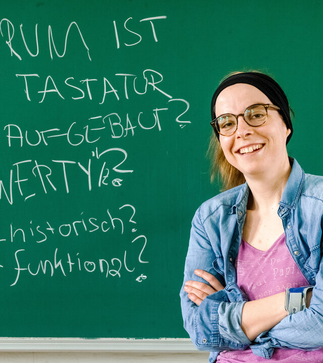 "einfach Mensch - Karola Köpferl: Inklusion an der Uni": Karola Köpferl sitzt im Rollstuhl in einem Hörsaal der TU Chemnitz vor einer grünen Tafel. Sie schaut lächelnd in die Kamera.