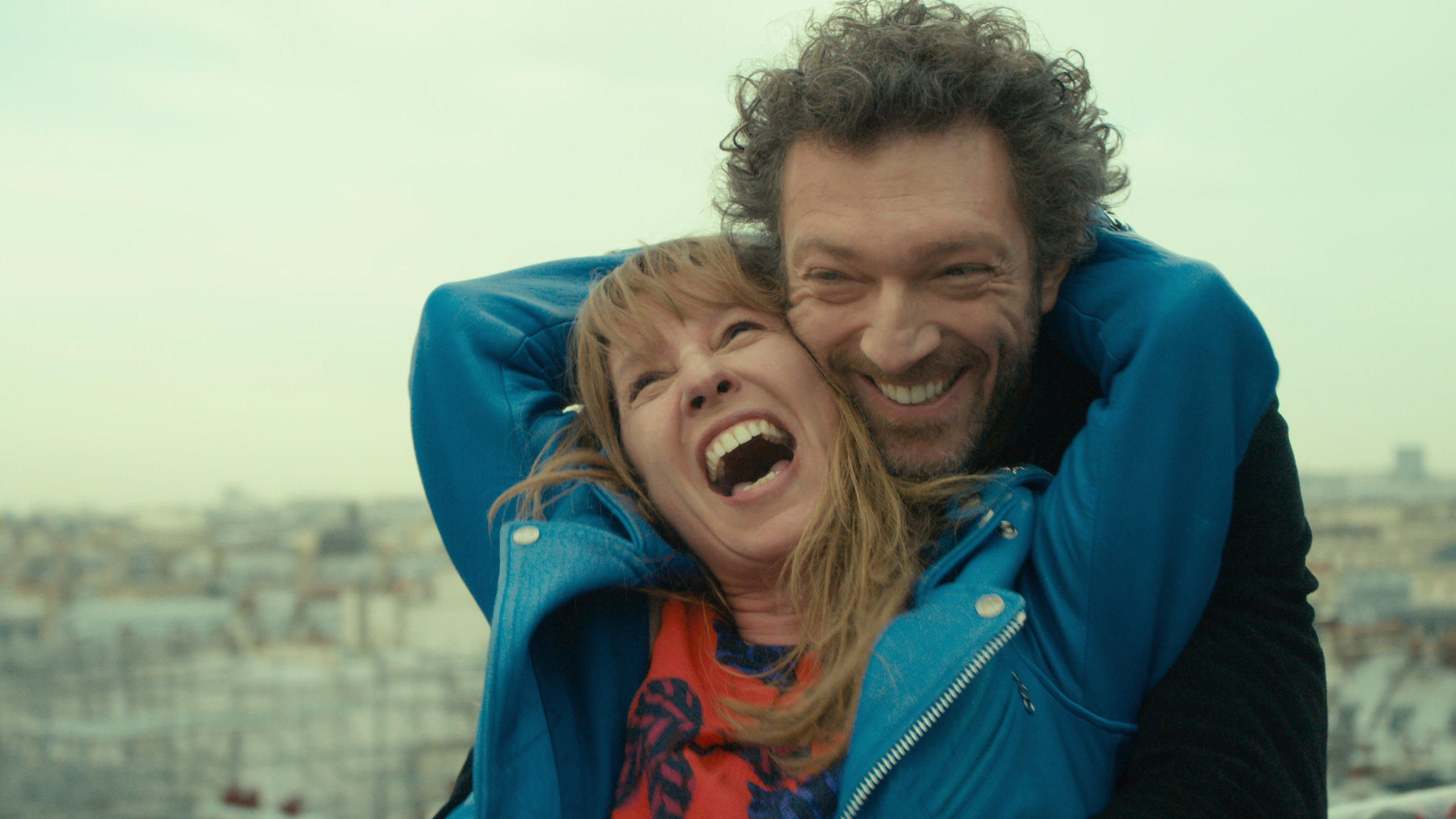 "Mein Ein, mein Alles": Giorgio (Vincent Cassel) steht lachend hinter seiner Geliebten Tony (Emanuelle Bercot), die glücklich die Arme um Giorgios Hals schlingt. Im Hintergrund sieht man verschwommen die Dächer einer Stadt.