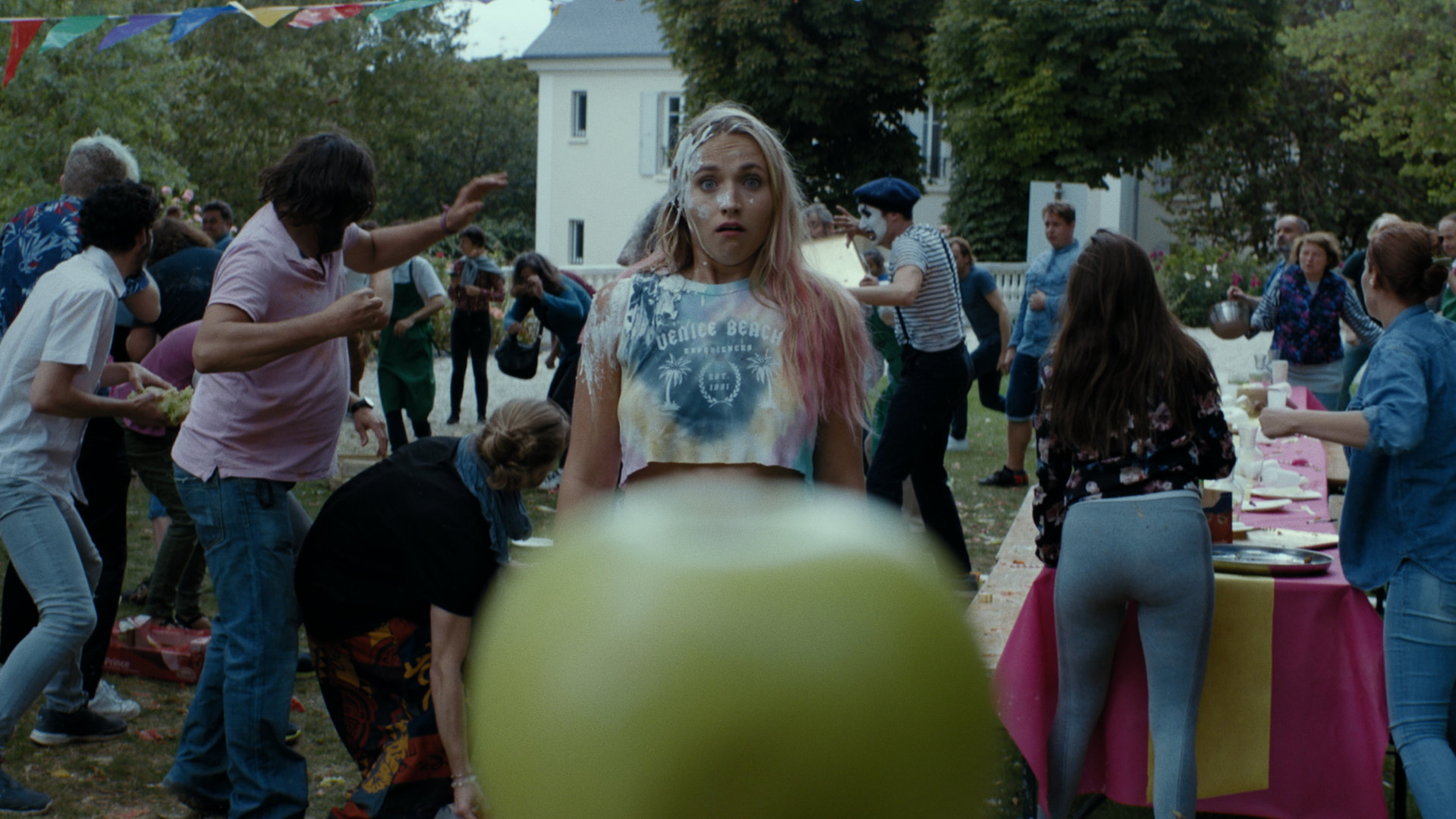 "Derby Girl - Tomatina!": Lola (Chloé Jouannet) steht zwischen zwei Menschenmengen, die sich im Hintergrund bekämpfen. Dabei schaut sie  mit einem mit Kuchen verschmierten Gesicht verstört nach vorne.