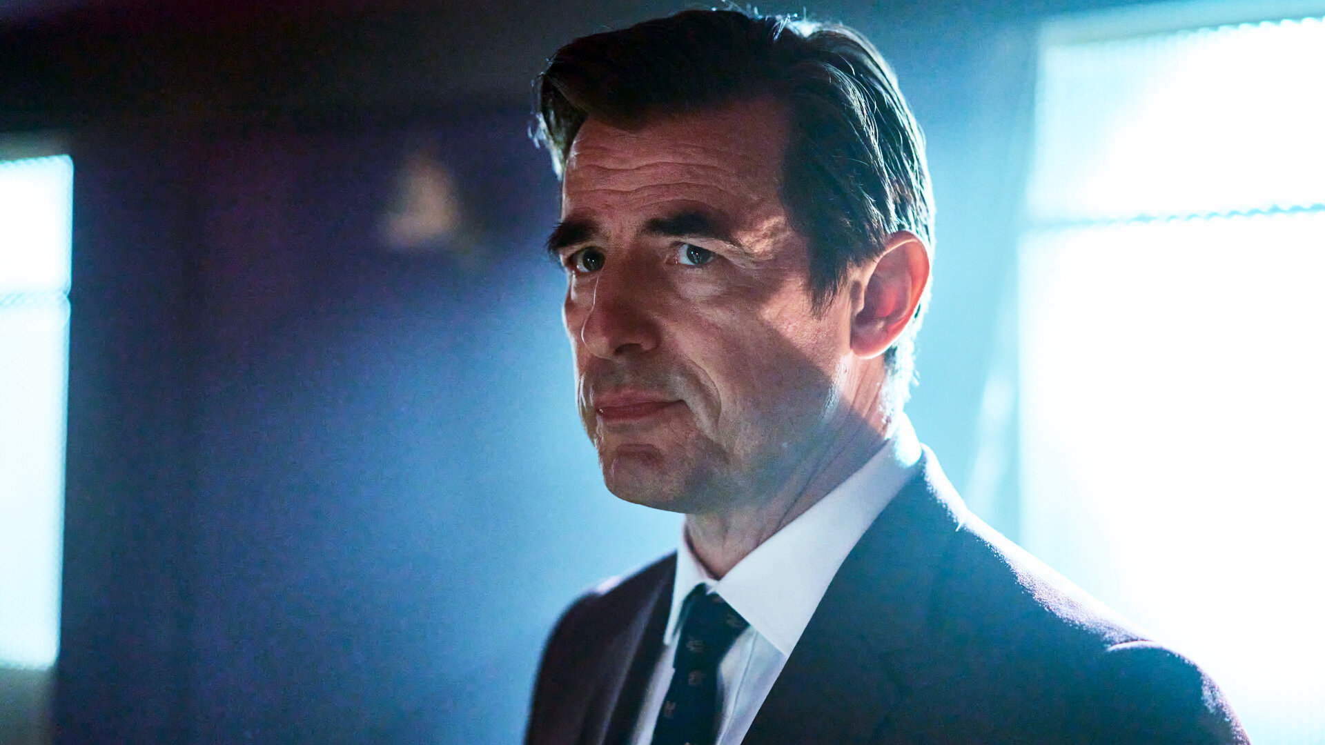 "The Outlaws - Es ist noch nicht vorbei": Der Dean (Claes Bang) steht in einem Raum mit dem Fenster im Rücken. Er trägt einen Anzug und schaut sehr ernst.