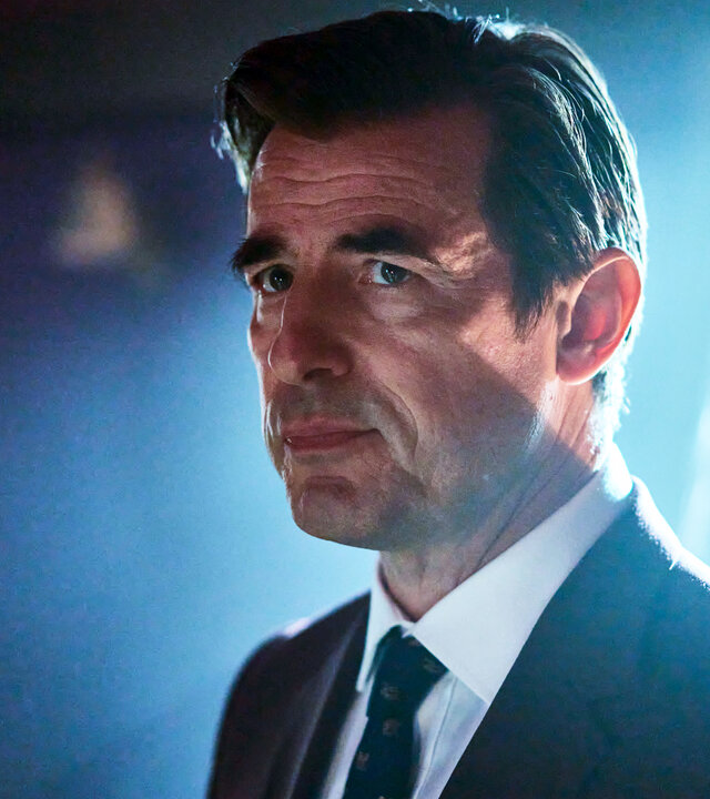 "The Outlaws - Es ist noch nicht vorbei": Der Dean (Claes Bang) steht in einem Raum mit dem Fenster im Rücken. Er trägt einen Anzug und schaut sehr ernst.